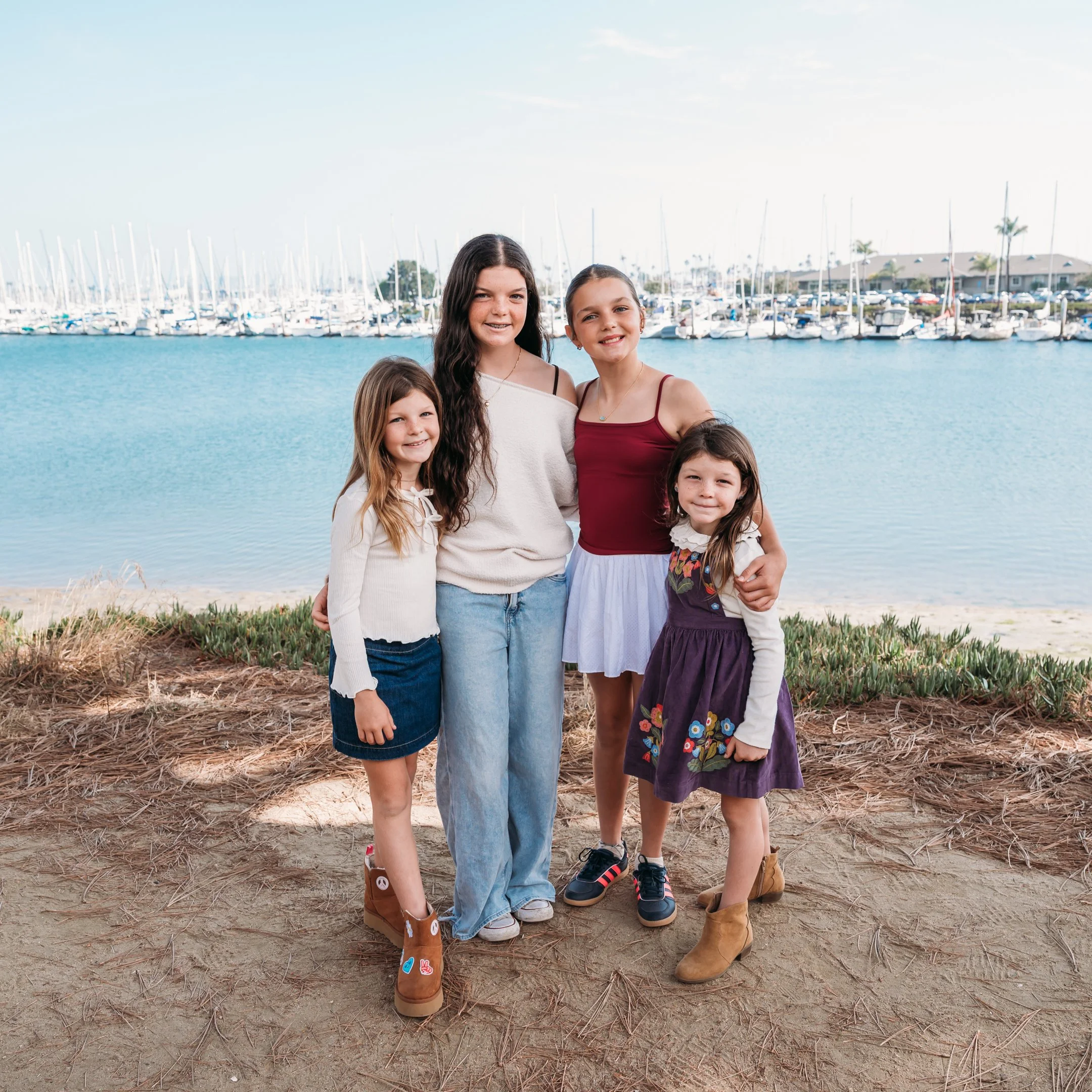Four young girls standing together near a marina with boats and a body of water in the background. Alisha Mowry Photography Military, Brand, and Portrait Photographer San Diego CA