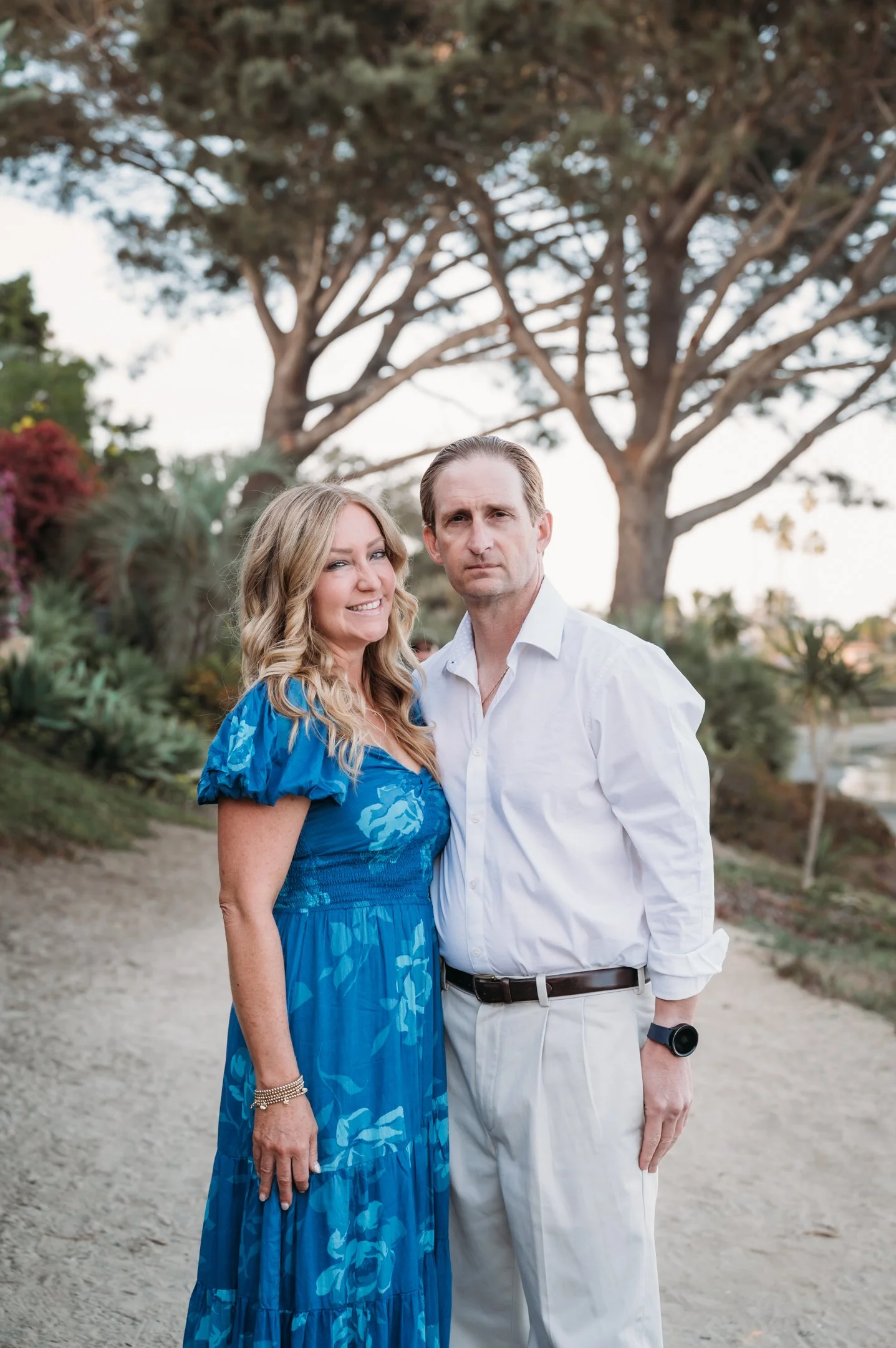 A woman in a blue dress with blonde hair and a man in a white shirt standing outdoors on a dirt path with trees and greenery behind them. Alisha Mowry Photography Military, Brand, and Portrait Photographer San Diego CA