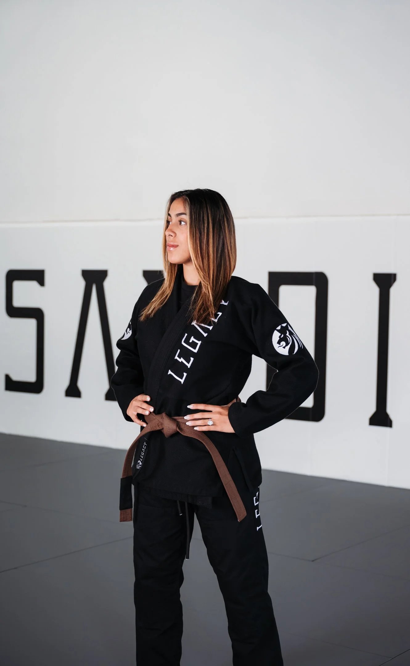 A woman in a black Brazilian jiu-jitsu gi with brown belt stands with hands on hips in a dojo, with the word 'LEGACY' visible on the gi and large letters spelling 'SAMBO' on the wall behind her. Alisha Mowry Photography Military, Brand, and Portrait 