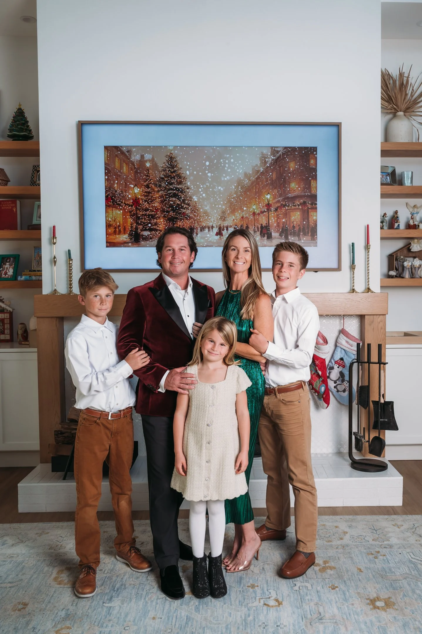 A family of five standing in front of a fireplace with holiday decorations inside a home. There are three boys and two girls, all dressed in semi-formal attire, smiling for a Christmas photo. The background features a large painting of a winter scene