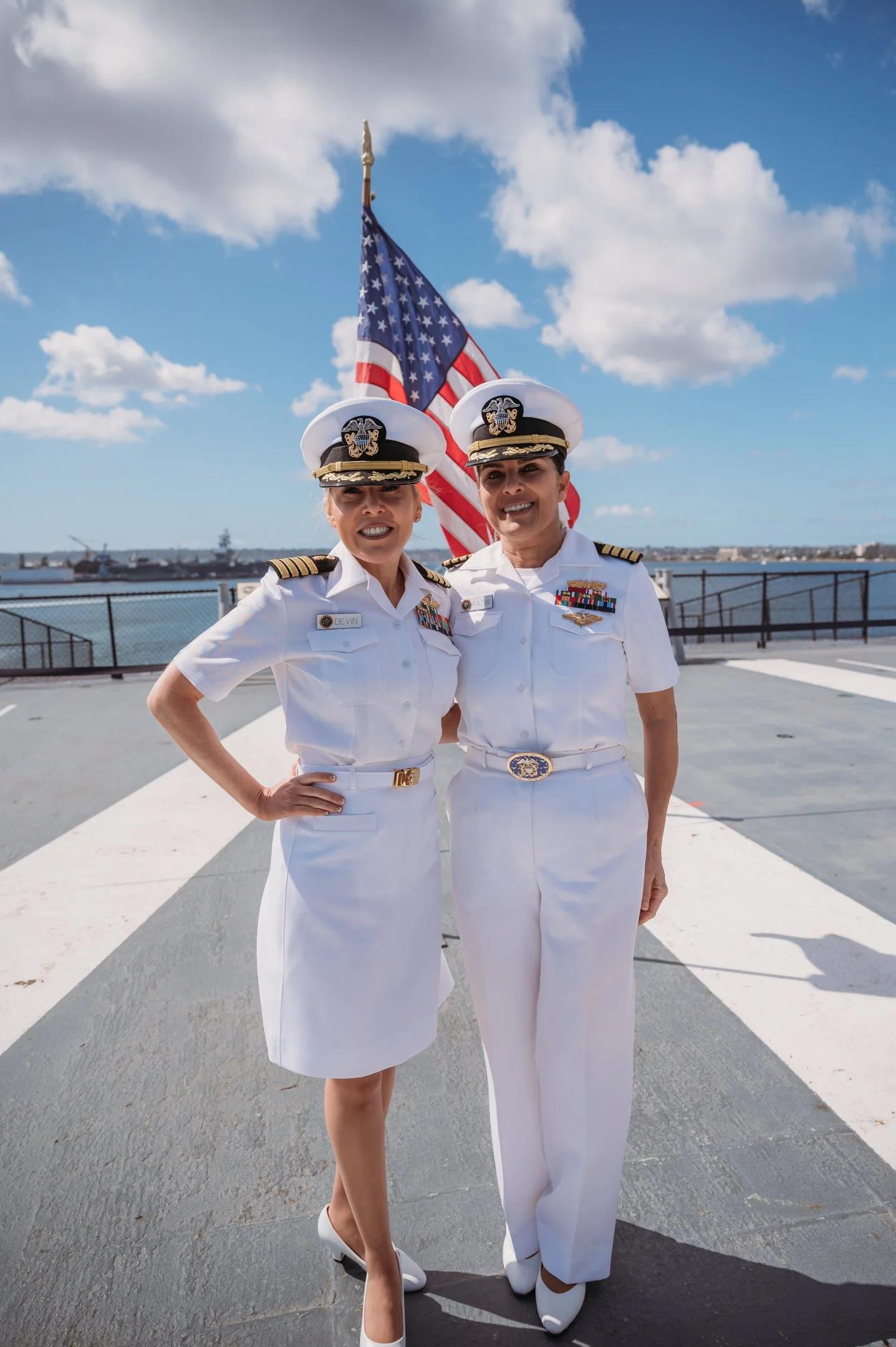 Two women in white military uniforms standing on a deck near a flagpole with the U.S. flag, with a body of water and ships in the background, under a partly cloudy sky. Alisha Mowry Photography Military Photographer San Diego
