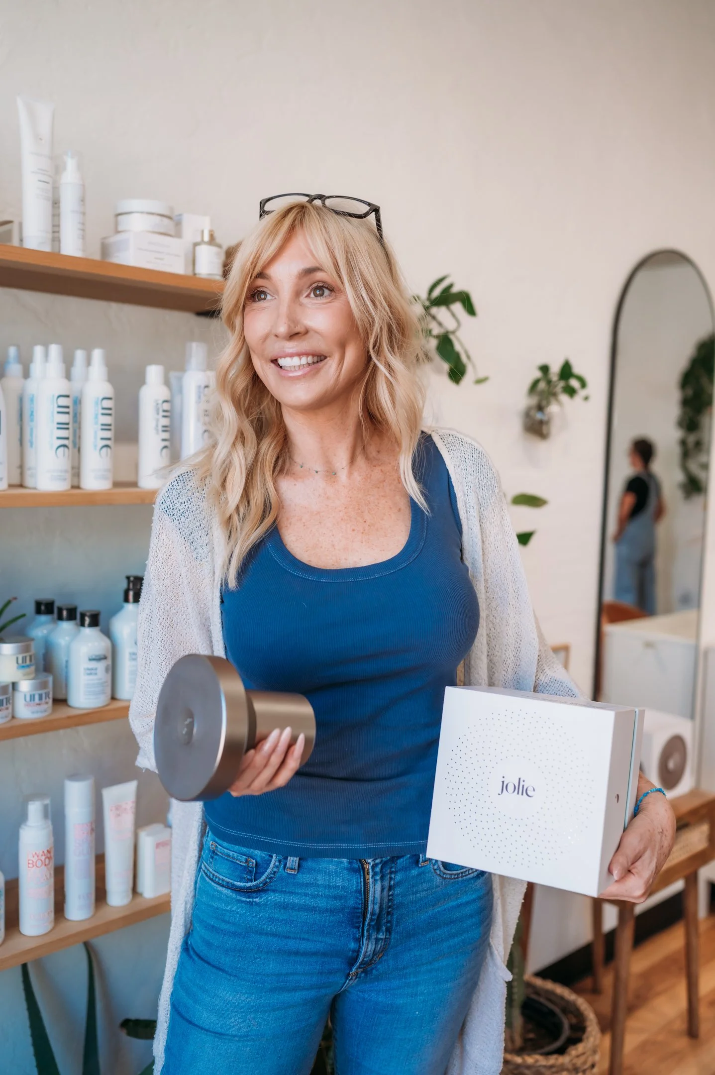 A woman with blonde hair holding a white box labeled 'Jolie' and a metal canister, standing inside a store with shelves of skincare products and a mirror in the background. Alisha Mowry Photography Military, Brand, and Portrait Photographer San Diego