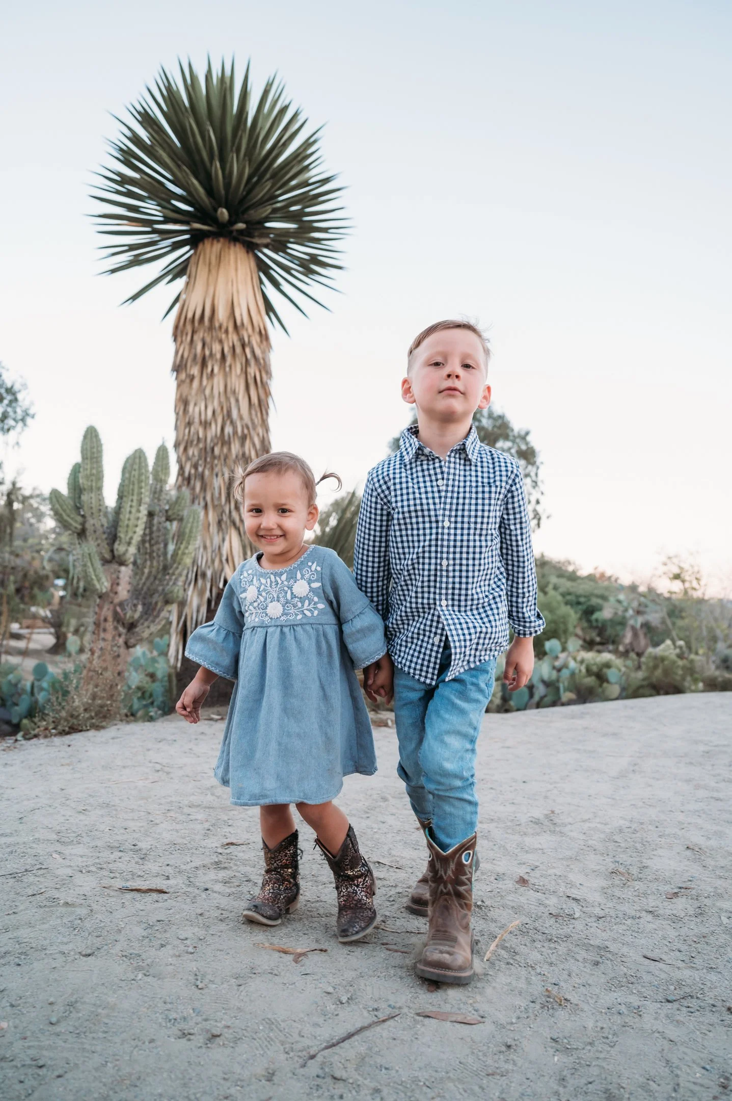 A young boy and girl walking hand in hand outdoors in a desert landscape with cactus and a tall palm tree, wearing casual clothing and boots. Alisha Mowry Photography Military, Brand, and Portrait Photographer San Diego CA