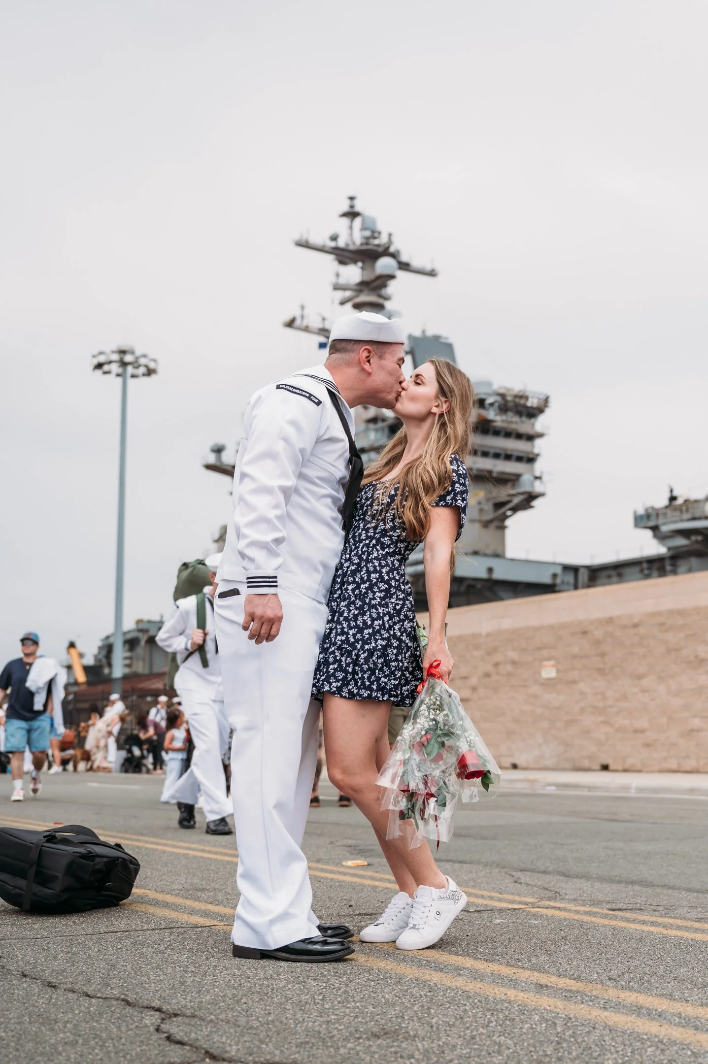 A couple kiss on a street in front of a large US aircraft carrier, with several people in the background.   San Diego Military Homecoming Photograph