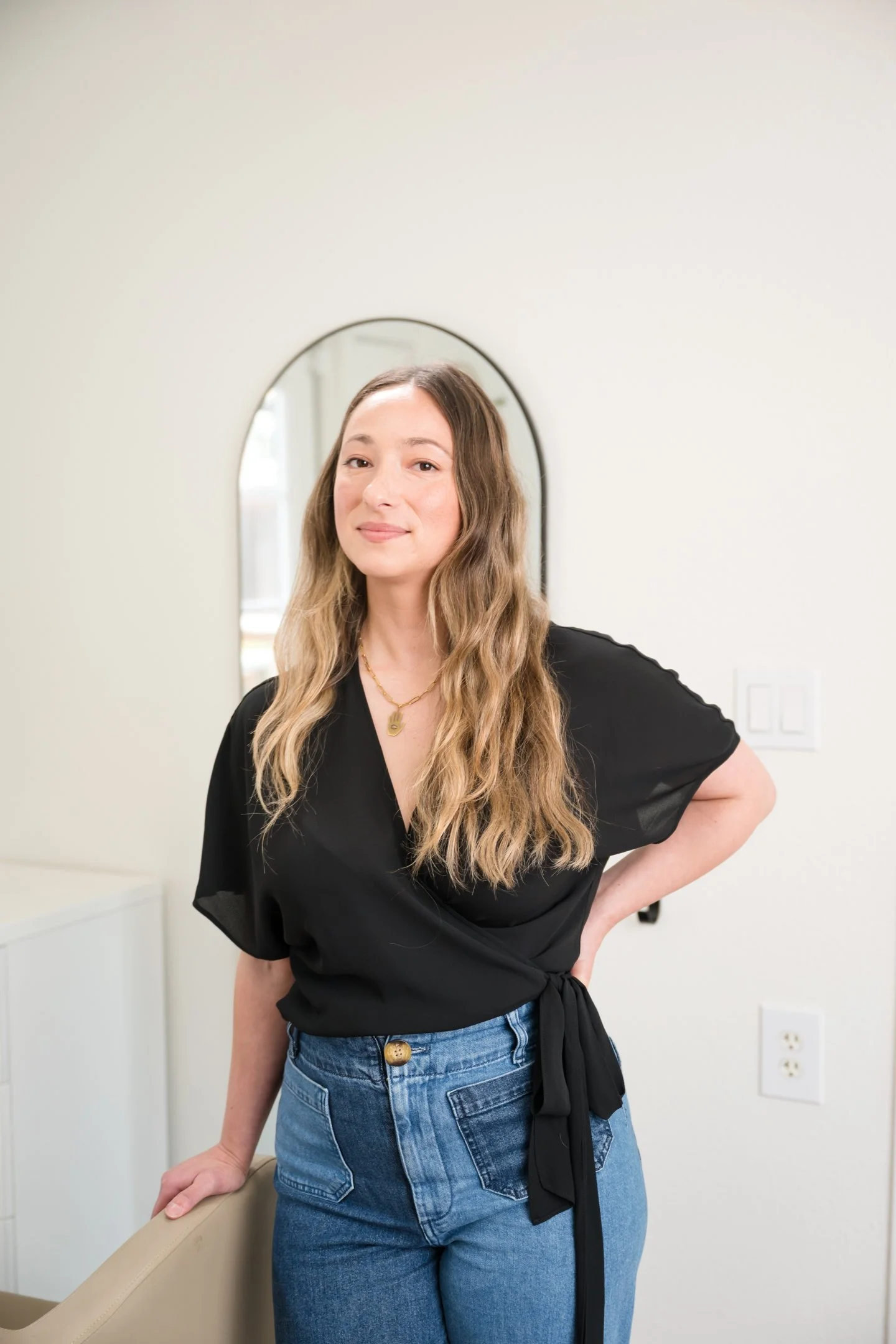 A woman with long wavy hair wearing a black top and blue jeans, standing in a room with a mirror and white walls. Alisha Mowry Photography Military, Brand, and Portrait Photographer San Diego CA