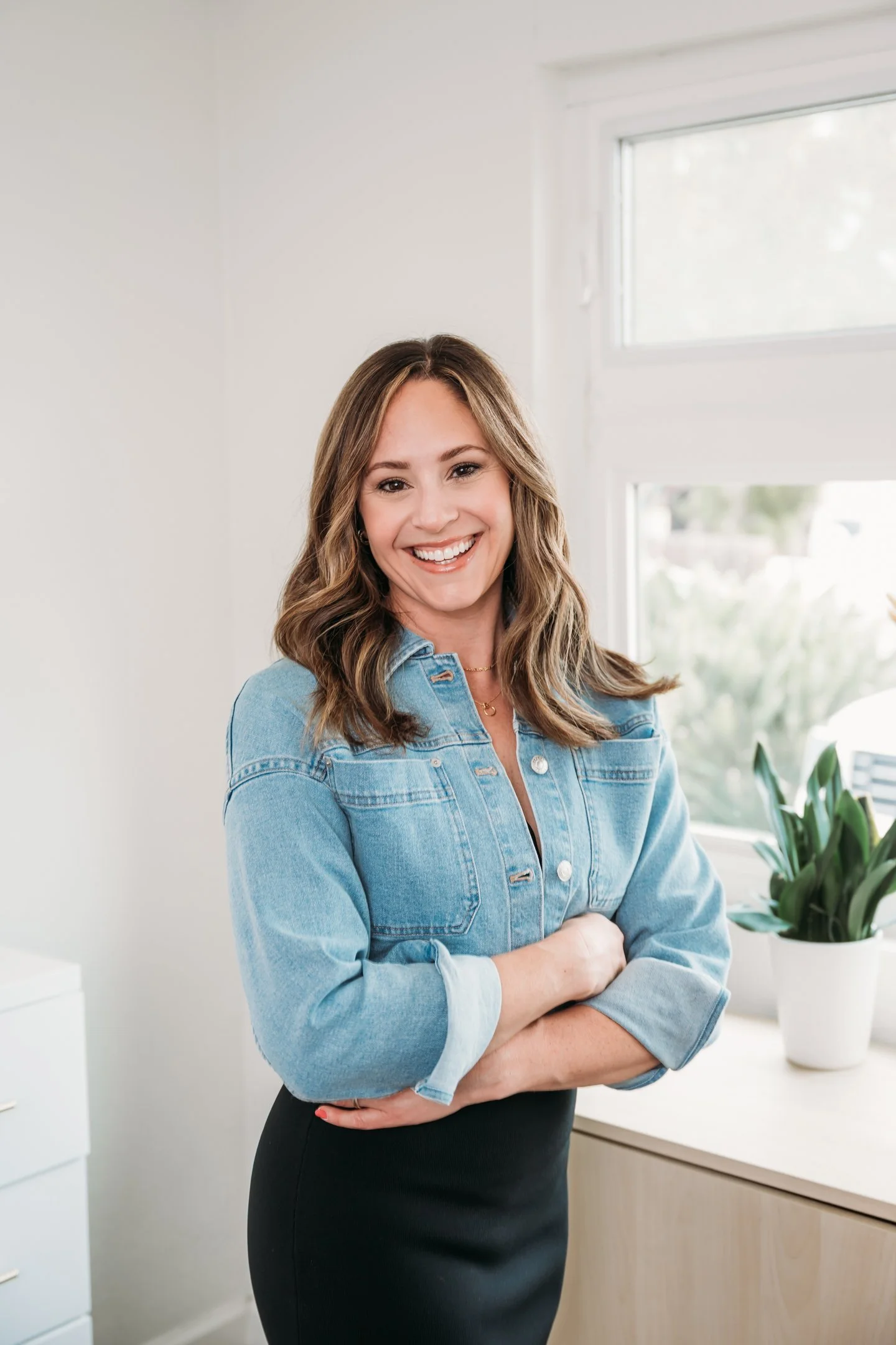 A woman with shoulder-length wavy brown hair and light skin smiling, standing with arms crossed in a bright room near a window, Alisha Mowry Photography Military, Brand, and Portrait Photographer San Diego CA