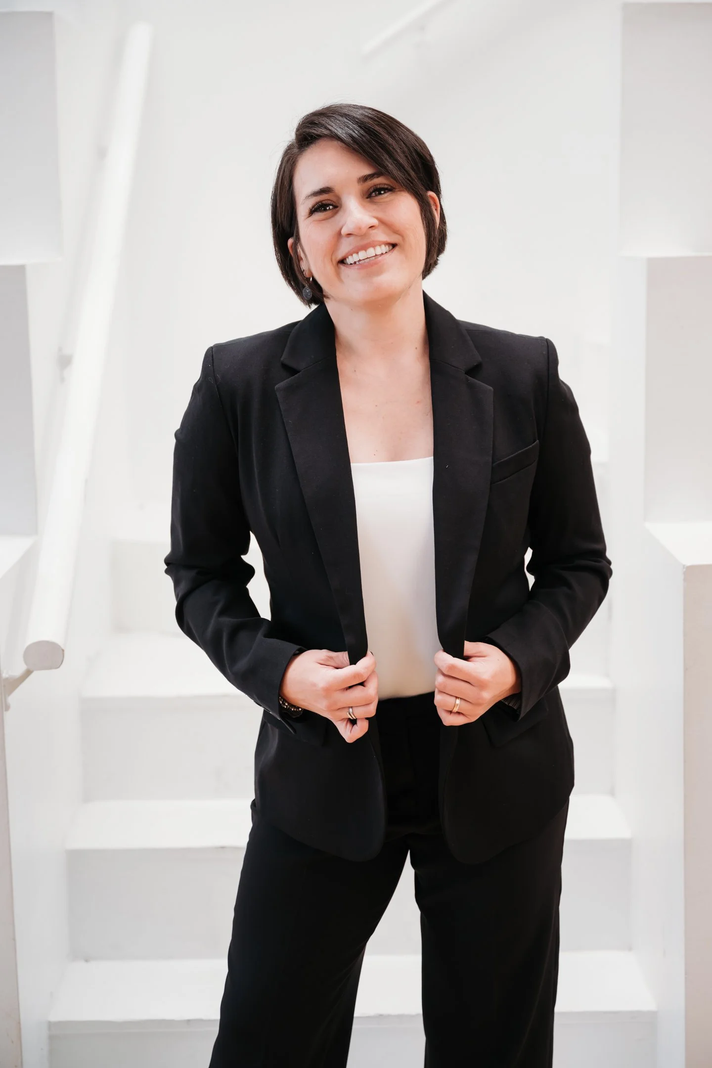 A woman with short dark hair smiling, wearing a black suit and white top, standing on white stairs indoors. Alisha Mowry Photography Military, Brand, and Portrait Photographer San Diego CA
