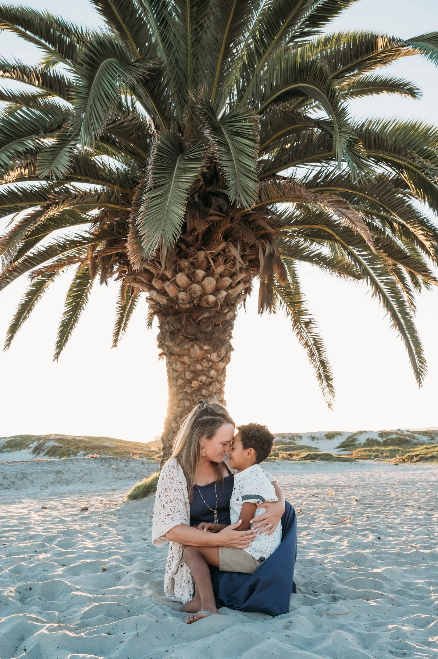 A woman and a young boy sitting on the sandy beach, embracing and touching foreheads under a large palm tree at sunset. Alisha Mowry Photography Military, Brand, and Portrait Photographer San Diego CA