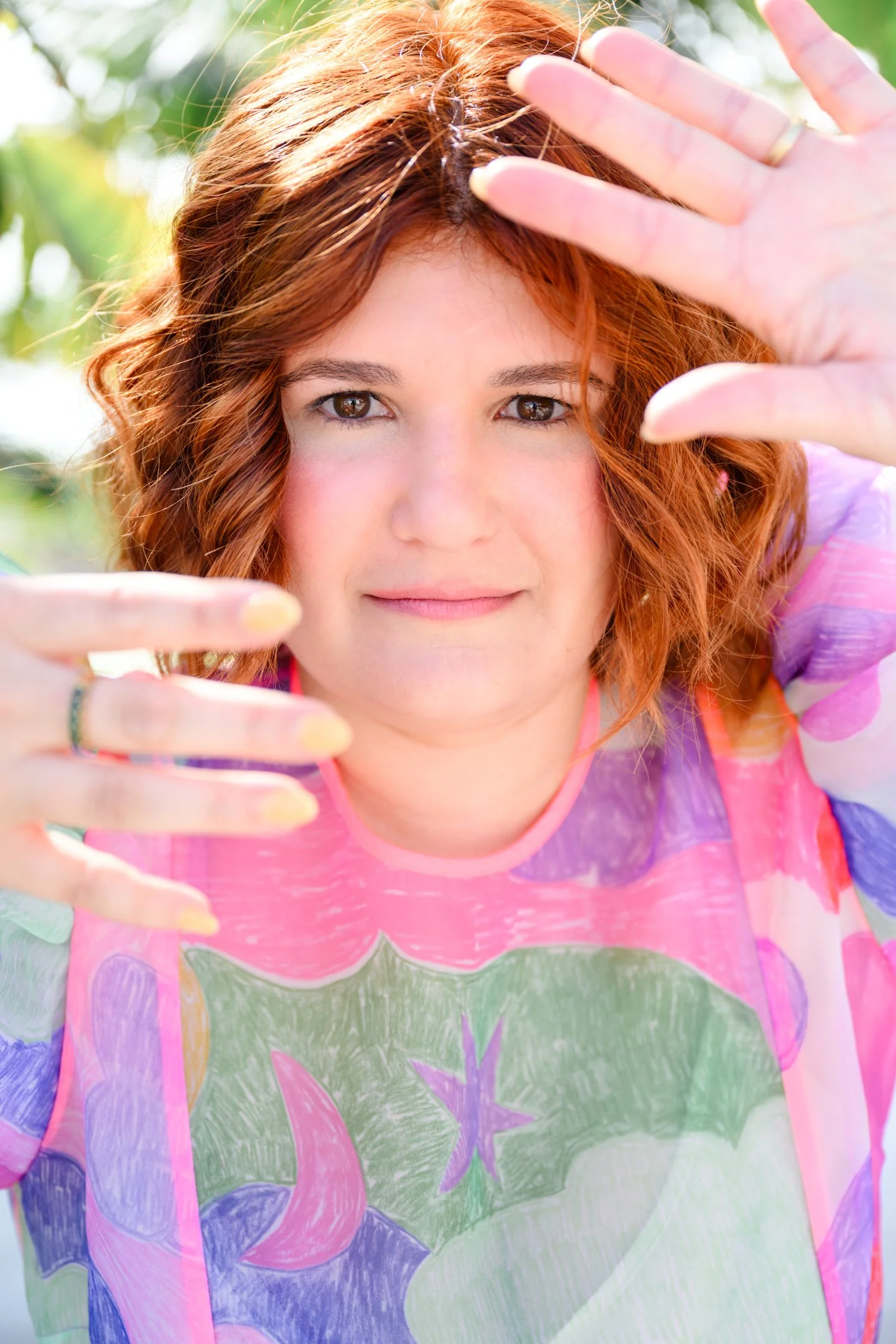 A woman with wavy red hair and light makeup holds her hands near her face, framing her face with her fingers, outdoors with greenery in the background. Alisha Mowry Photography Military, Brand, and Portrait Photographer San Diego CA