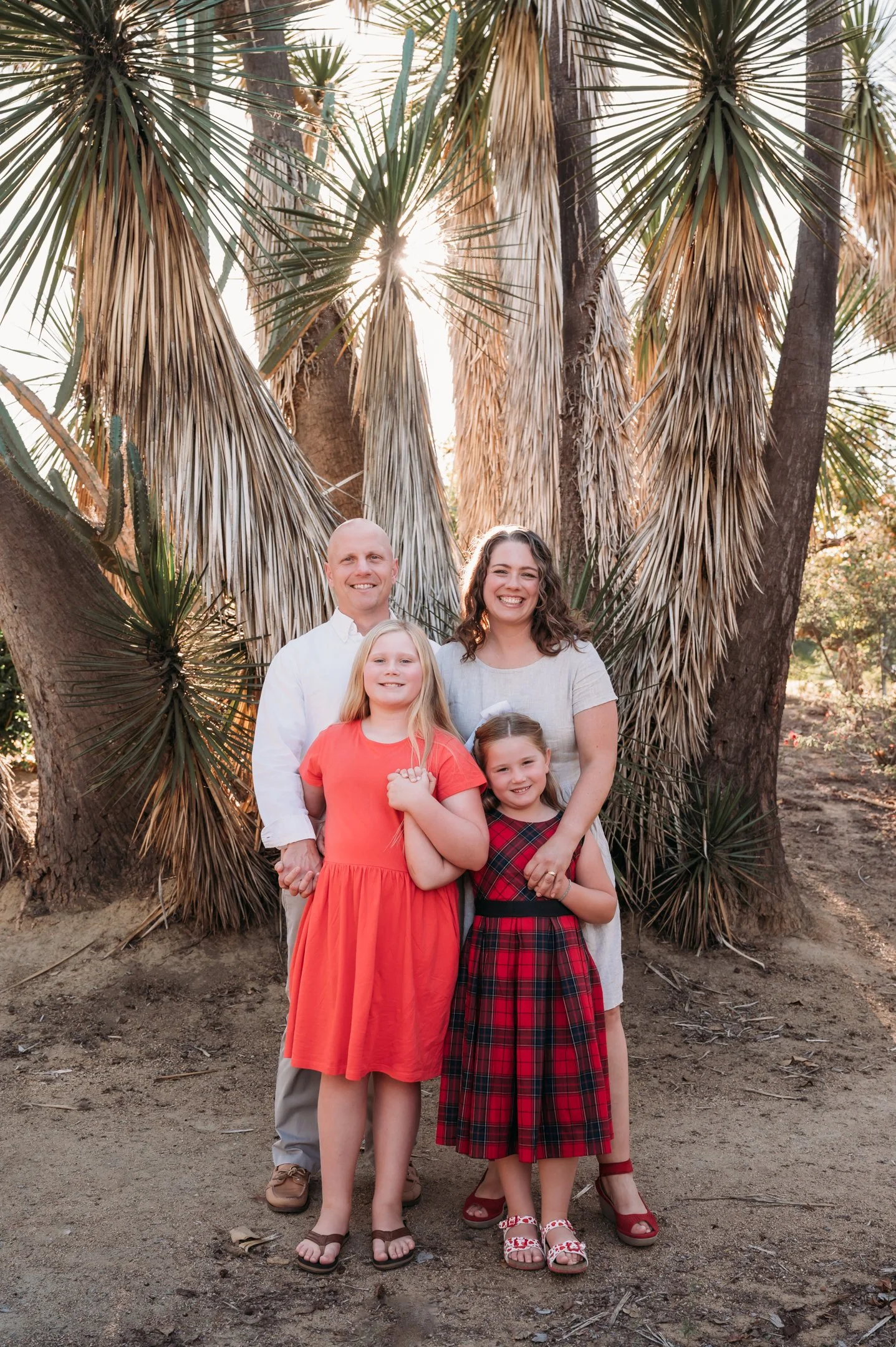 A family of four standing outdoors in front of palm trees during sunset. The father and mother are smiling, with the mother and two young daughters in dresses, one in orange and the other in red plaid. Alisha Mowry Photography Military, Brand, and Po