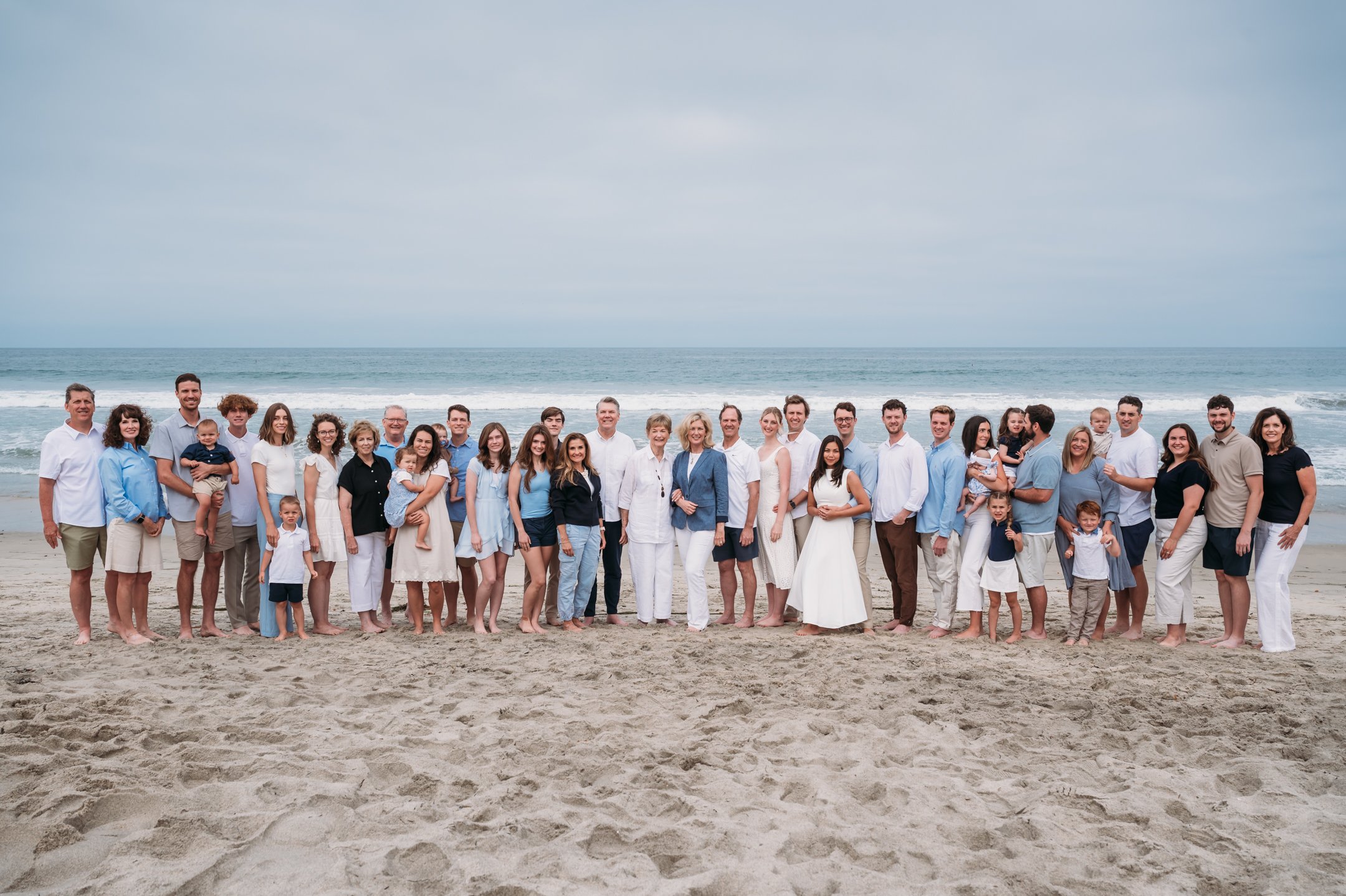 Large group of people including children and adults standing on a sandy beach with the ocean and sky in the background.