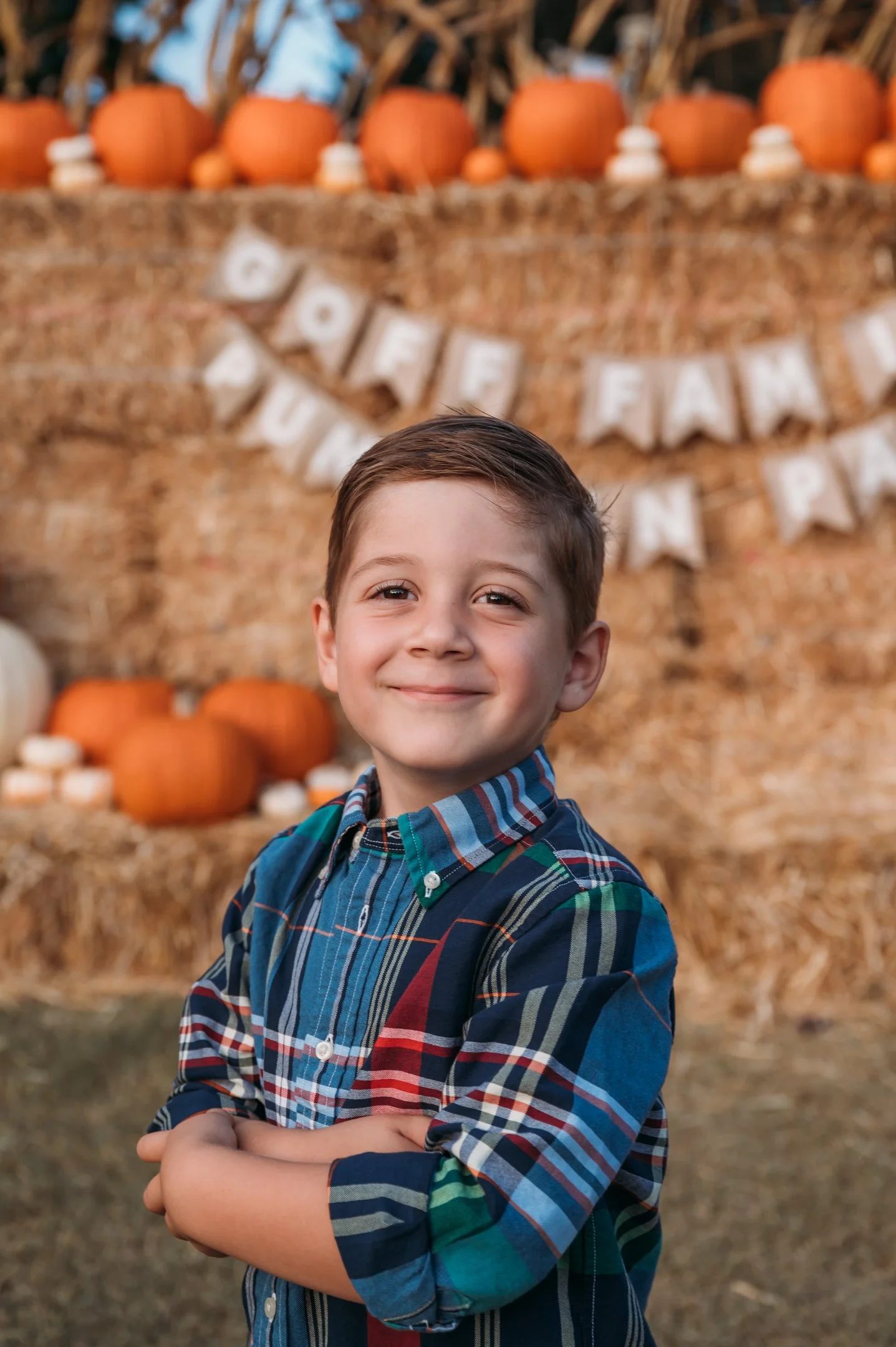A young boy smiling with arms crossed at a pumpkin patch decorated for Halloween with pumpkins, hay bales, and a 'GOBBLE, PLUCK, PASS' banner. Alisha Mowry Photography Military, Brand, and Portrait Photographer San Diego CA