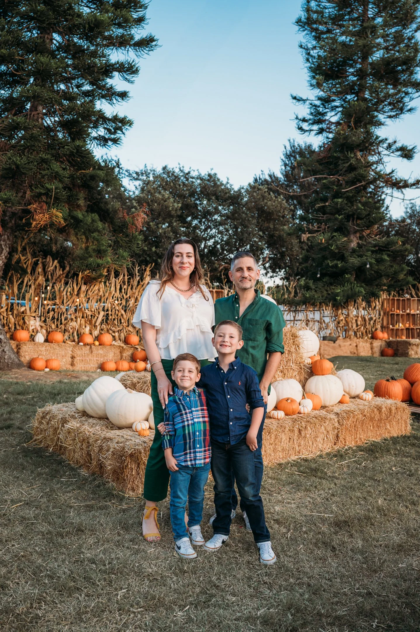 Family of four posing outdoors during fall. Behind them are pumpkins, hay bales, and corn stalks, with tall trees and a clear sky in the background. Alisha Mowry Photography Military, Brand, and Portrait Photographer San Diego CA