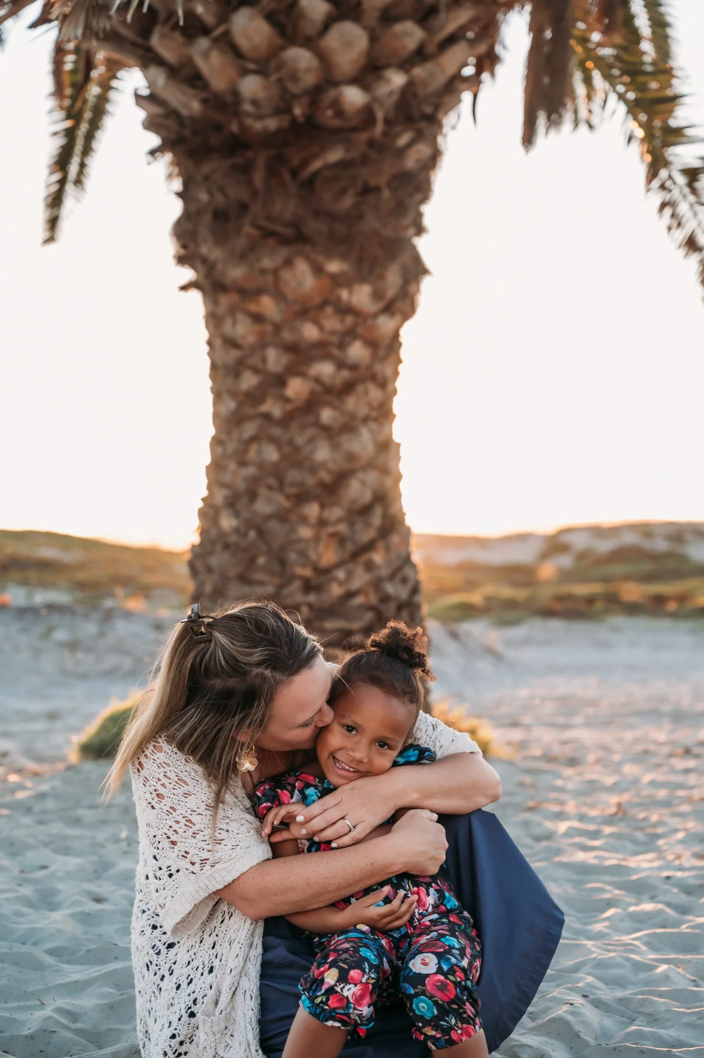 A woman and a young girl hugging on a sandy beach with a palm tree and sunset in the background. Alisha Mowry Photography Military, Brand, and Portrait Photographer San Diego CA
