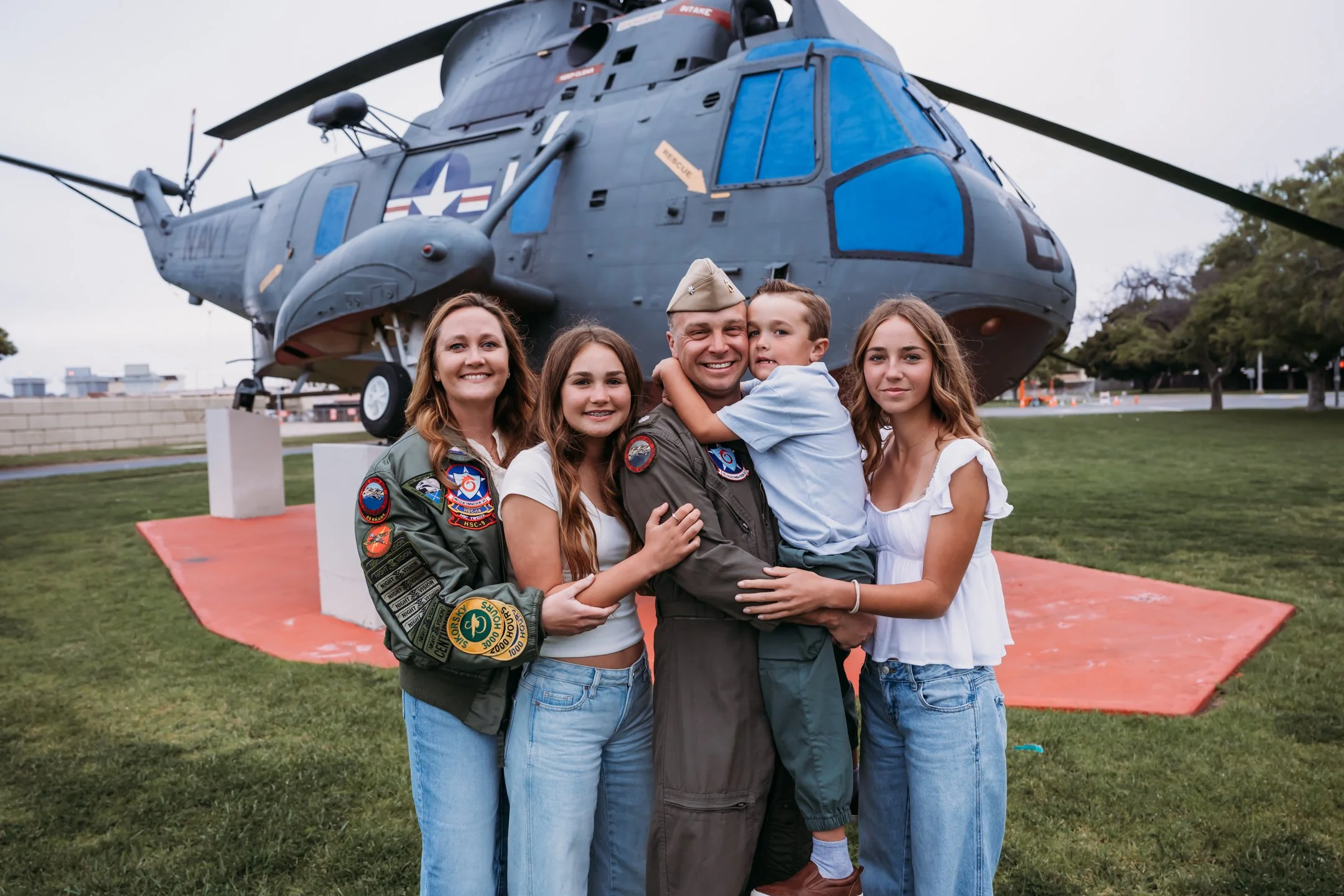 A smiling family of five hugging in front of a large military helicopter on display outdoors, with grassy area and trees in the background.