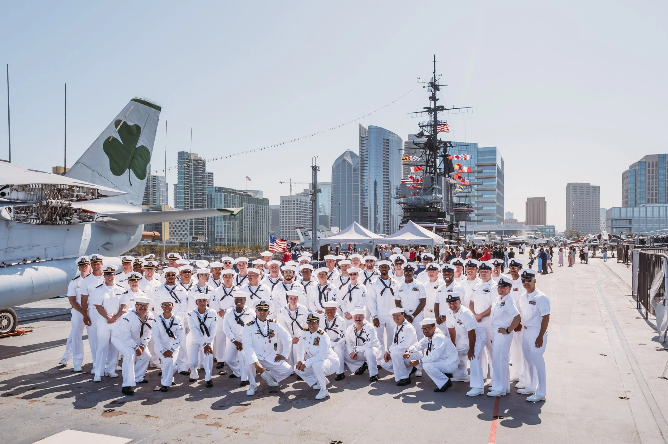 Group of sailors in white uniforms posing on the deck of a navy ship at an air and space museum with various aircraft and city skyscrapers in the background. Alisha Mowry Photography Military Photographer San Diego