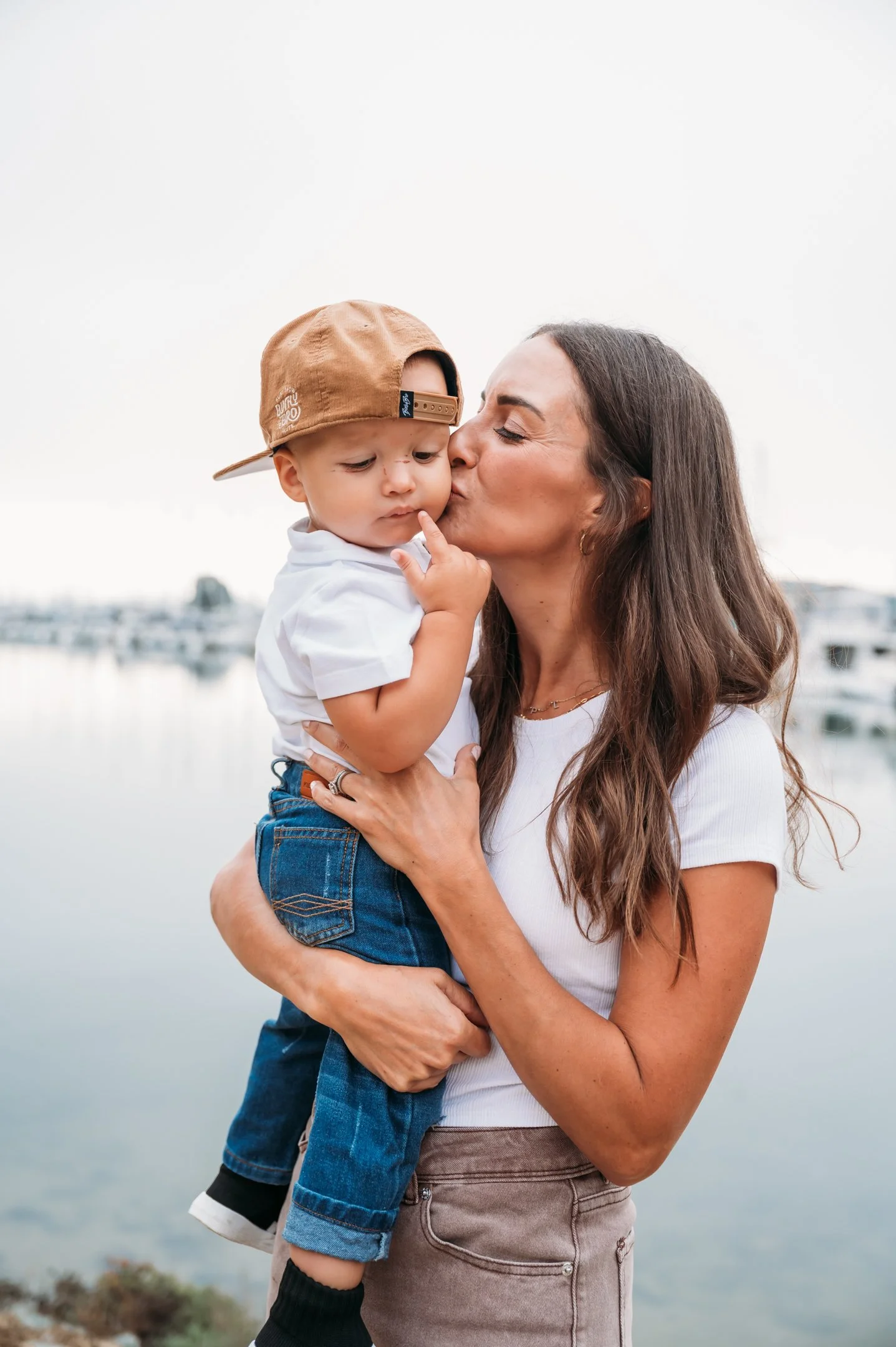 A woman holding a young boy by a waterfront with boats in the background, the woman is kissing the child's cheek.