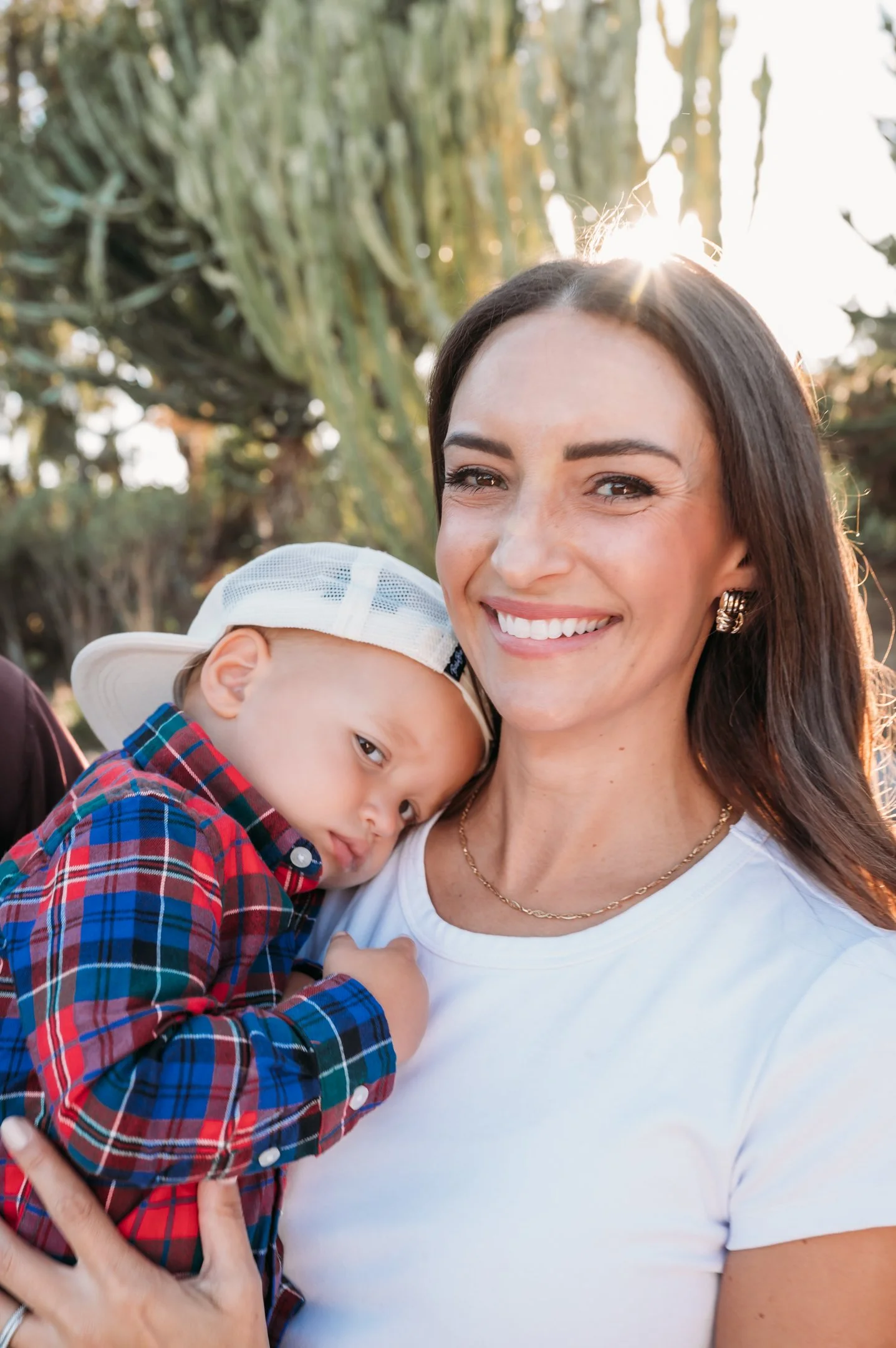 A smiling woman holding a young boy with his head resting on her shoulder, outdoors with trees and sunlight shining through in the background. Alisha Mowry Photography Military, Brand, and Portrait Photographer San Diego CA