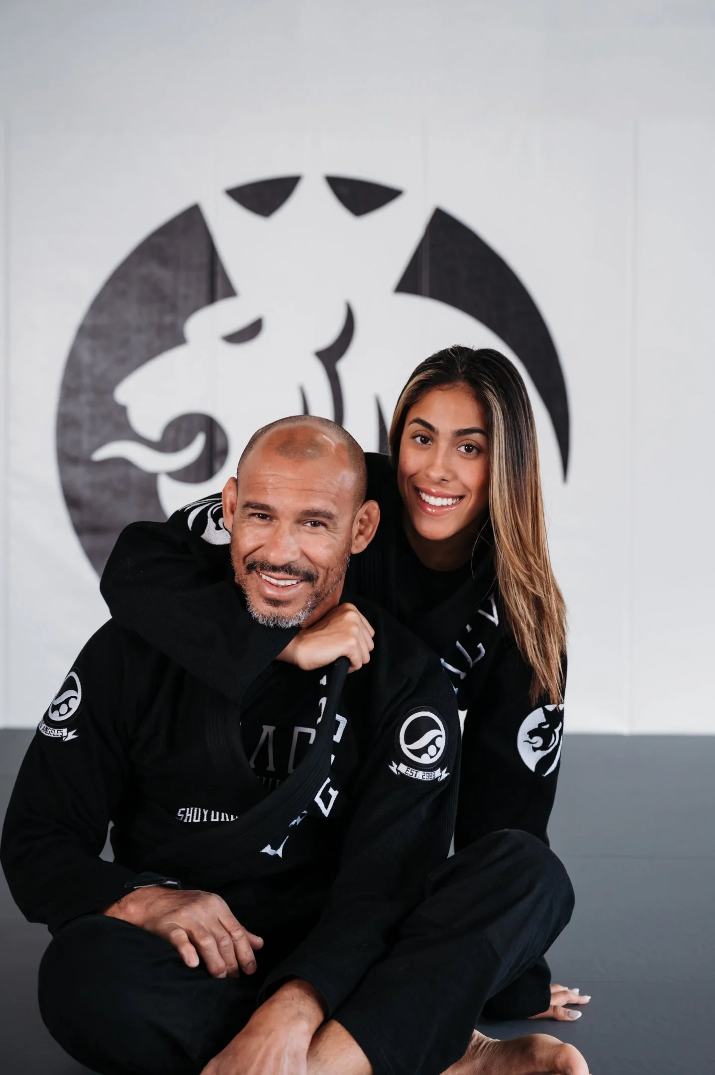 Two martial artists in black uniforms smiling, one with a woman on his back, in a gym with a logo of a panther on the wall. Alisha Mowry Photography Military, Brand, and Portrait Photographer San Diego CA