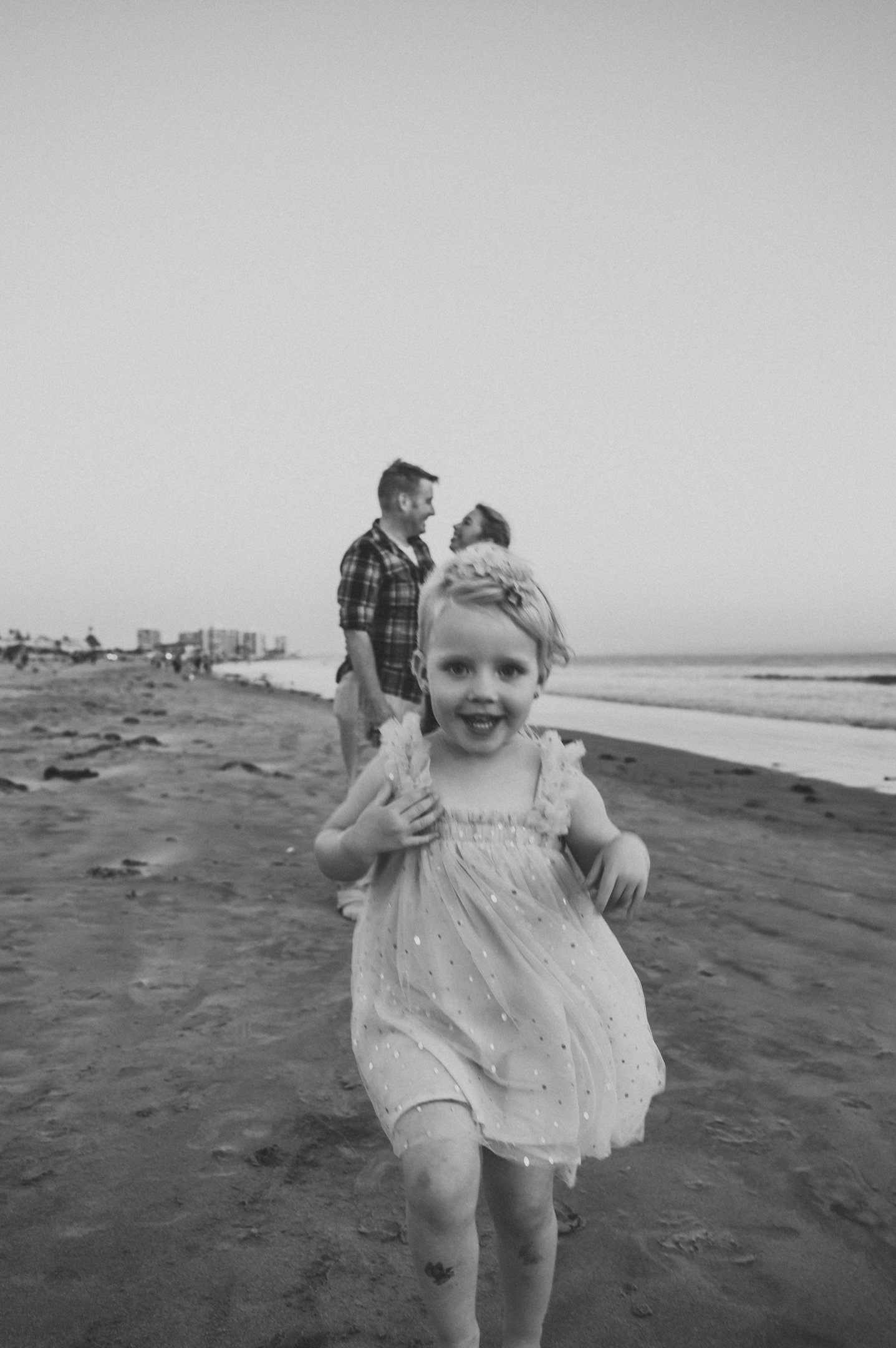 A young girl running on the beach towards the camera, smiling, with two adults in the background and a city skyline in the distance. Alisha Mowry Photography Military, Brand, and Portrait Photographer San Diego CA