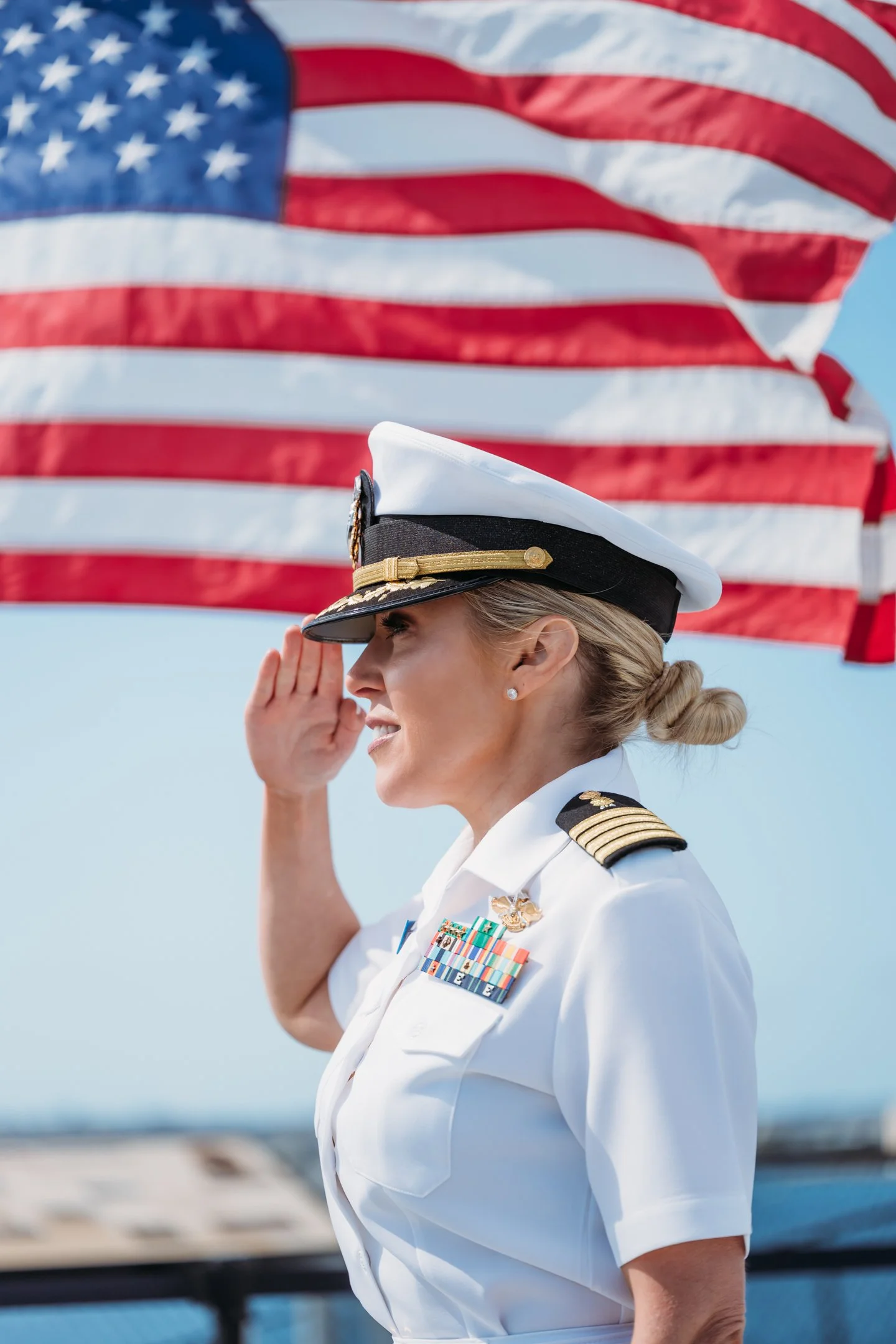 A woman in a white military uniform salutes with her right hand as she stands in front of a waving American flag. uss midway military promotion ceremony 