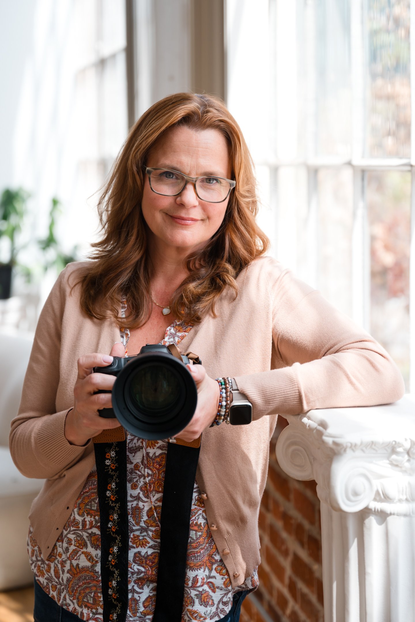 A woman standing indoors near large windows, holding a camera, smiling, with a potted plant in the background, wearing glasses, a beige cardigan, and a patterned blouse. Alisha Mowry Photography Military, Brand, and Portrait Photographer San Diego CA