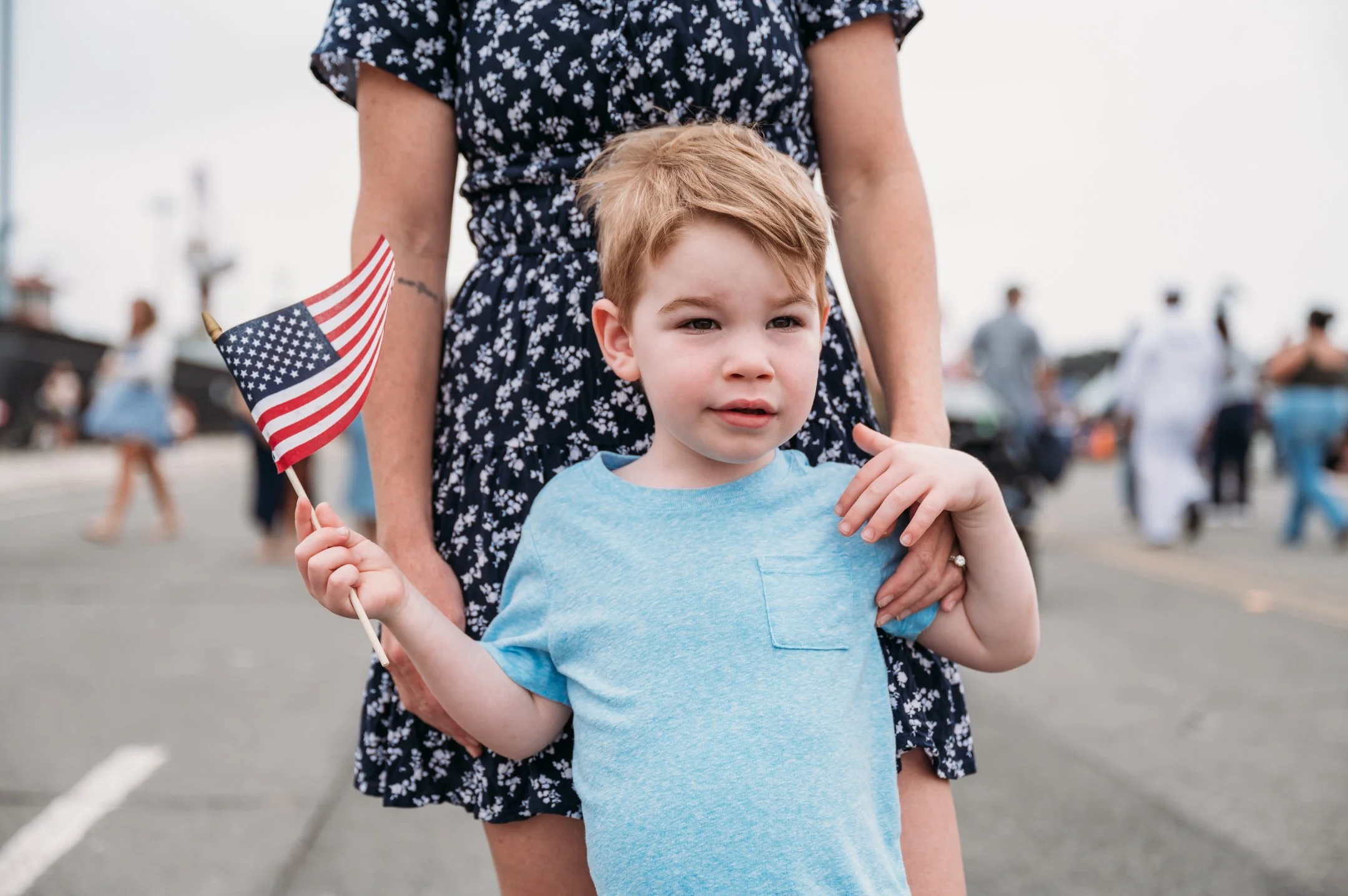 A young boy with red hair holding an American flag in his right hand while standing in front of a woman at an outdoor event with blurred people in the background.   San Diego Military Homecoming Photograph