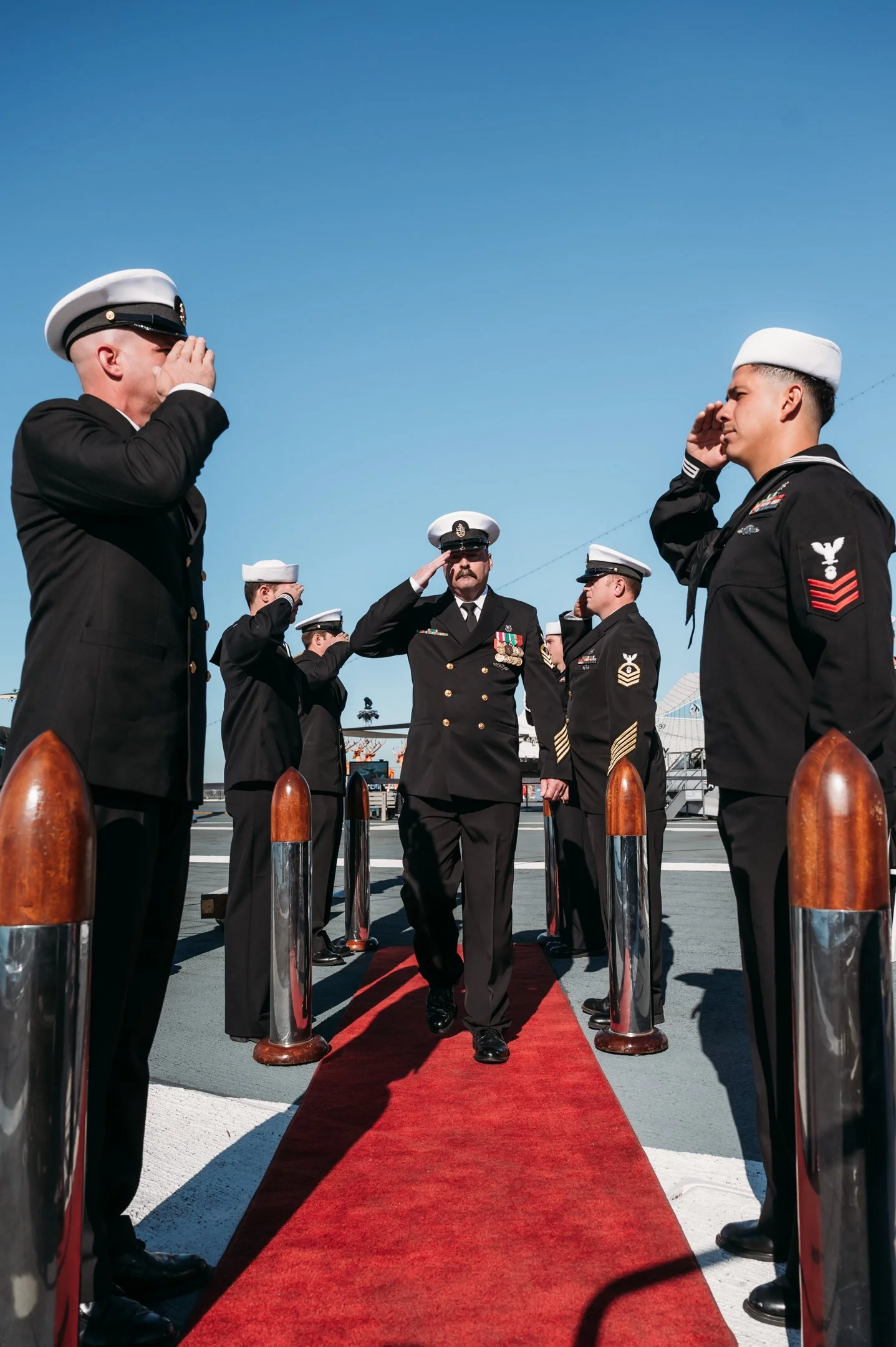 A military officer walking on a red carpet while saluting, surrounded by personnel in uniform saluting and saluting back at an outdoor event on the deck of a ship under a clear blue sky. uss midway military retirement ceremony 