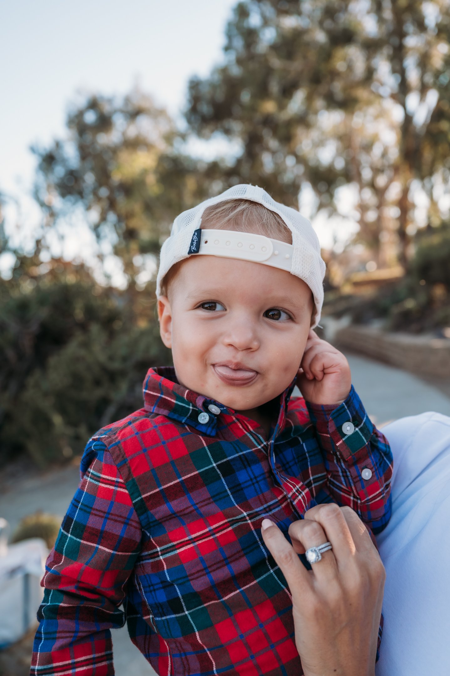 A young boy with light brown hair, wearing a white cap backward and a red and blue plaid shirt, sticking out his tongue while being held by someone with a wedding ring, outdoors with trees in the background. Alisha Mowry Photography Military, Brand, 
