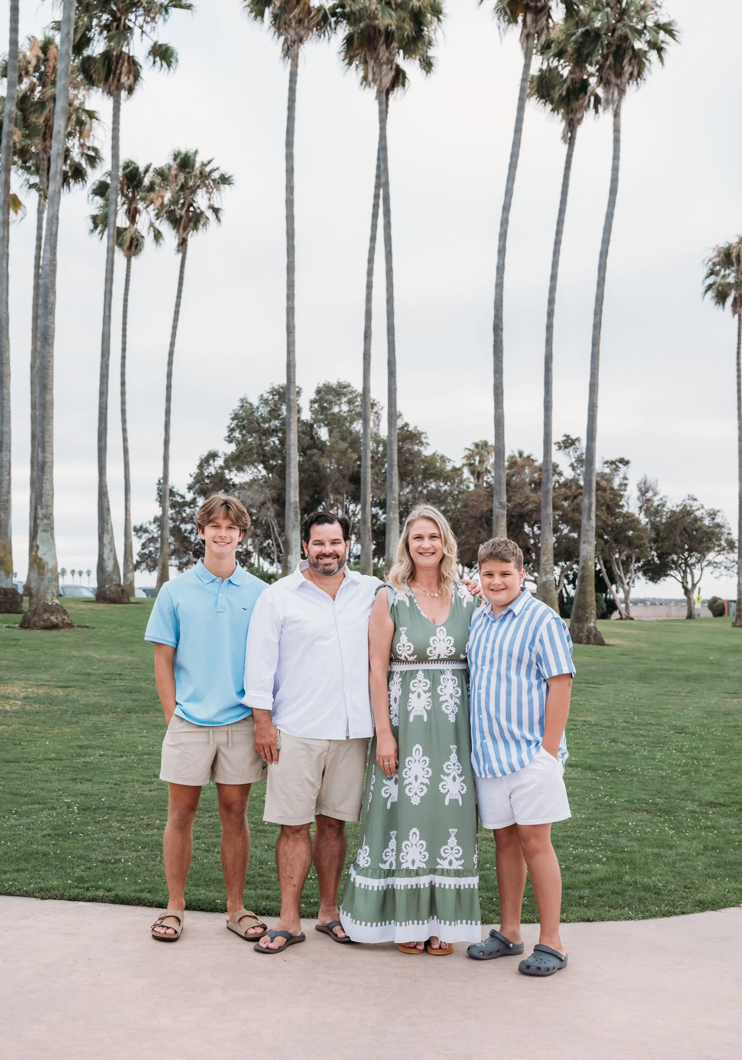 Family of four standing outdoors on a grassy area with tall palm trees and cloudy sky in the background.