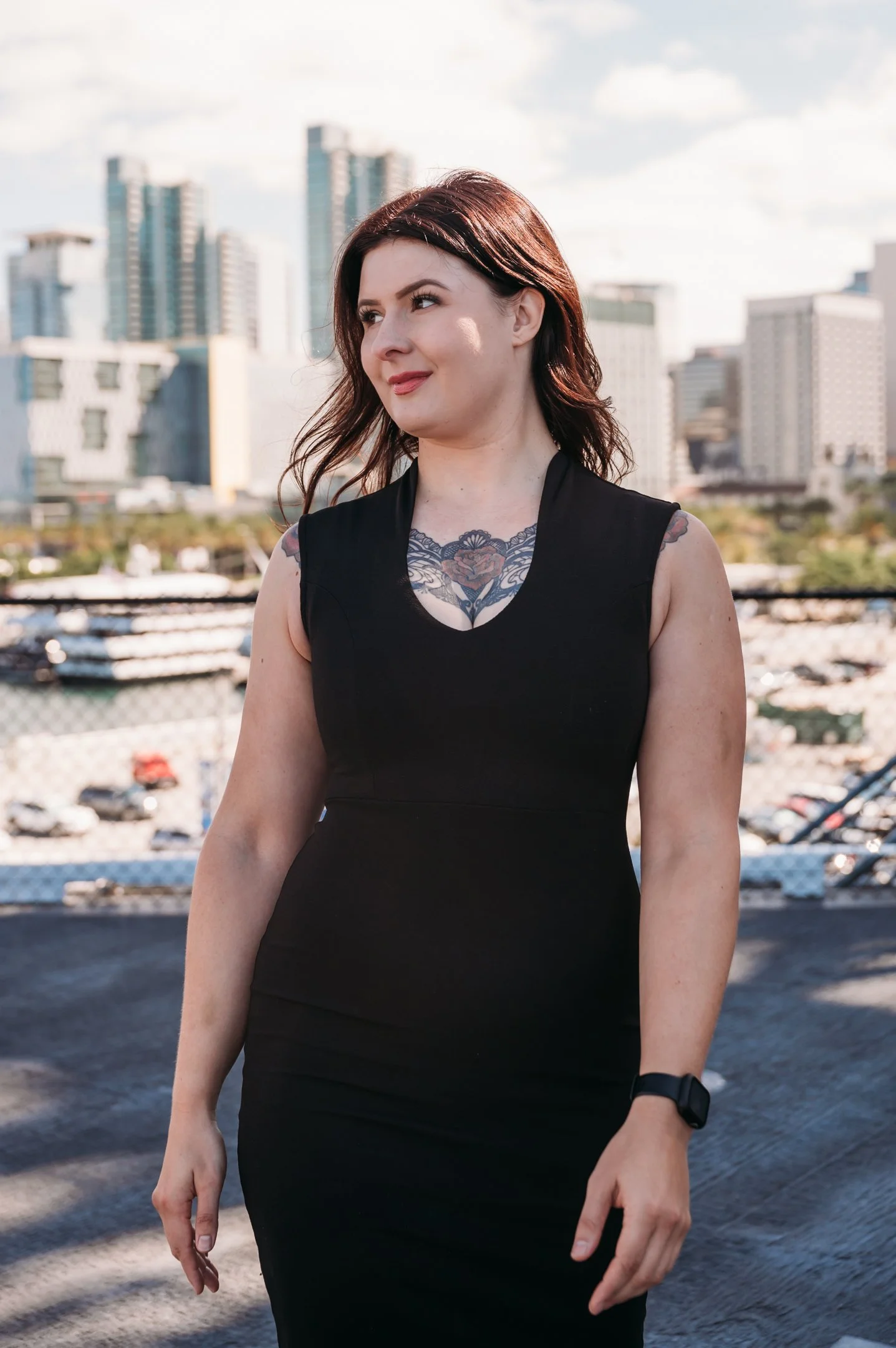 A woman with shoulder-length brown hair, wearing a black sleeveless dress and a watch, stands outdoors in an urban setting with tall buildings and a marina in the background. Alisha Mowry Photography Military, Brand, and Portrait Photographer San Die