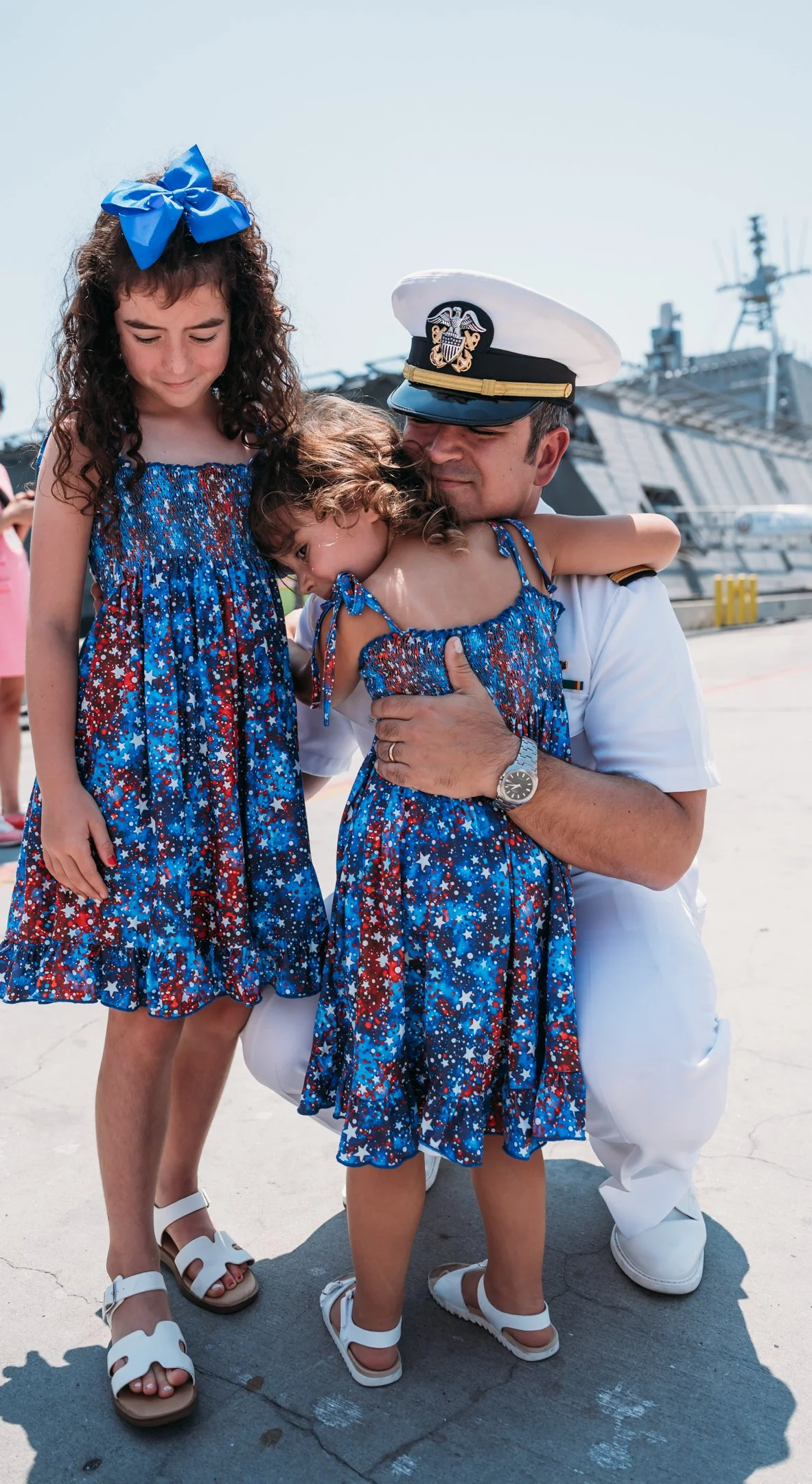 A sailor in uniform hugging two young girls in patriotic dresses with star prints, at a dock with ships in the background.