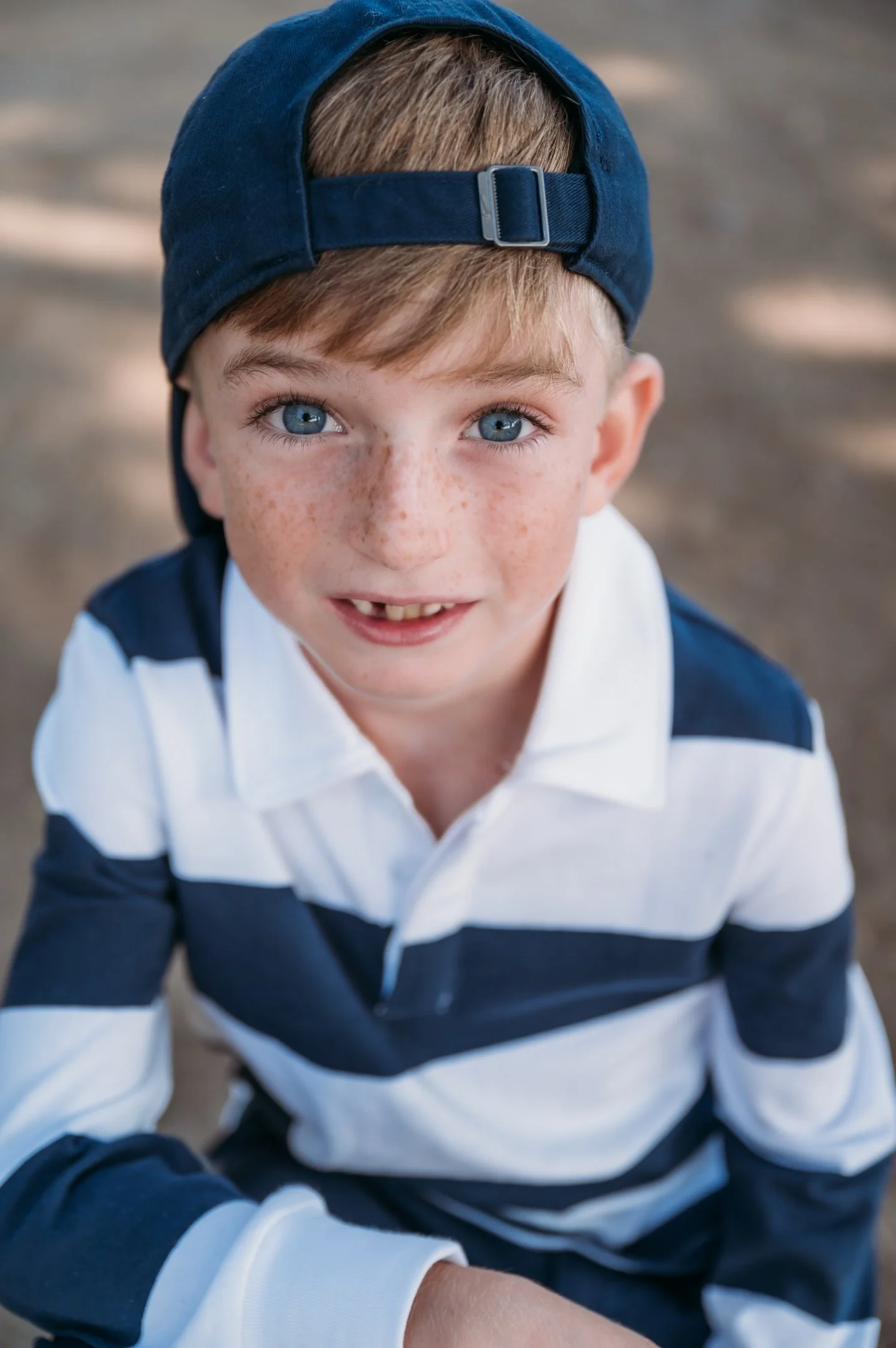 Close-up of a young boy with blue eyes, freckles, and missing front teeth, wearing a navy blue cap backwards and a navy and white striped shirt, smiling outdoors. Alisha Mowry Photography Military, Brand, and Portrait Photographer San Diego CA