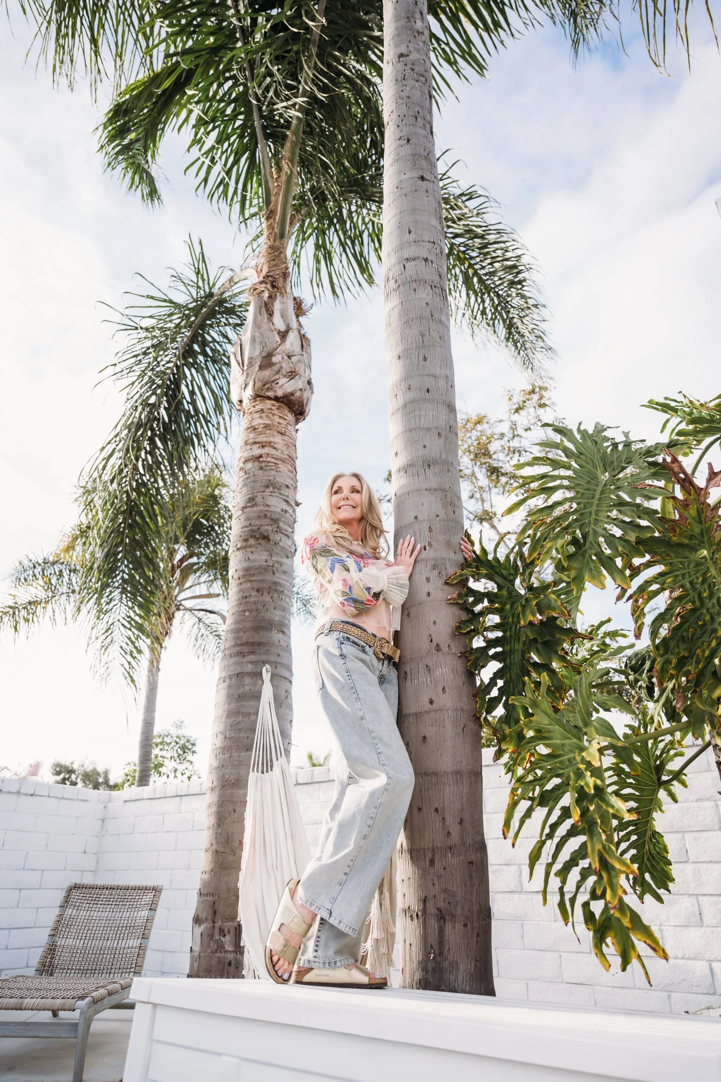 A woman in casual clothing and sandals is standing on a white platform, hugging a palm tree, with another palm tree and a leafy plant in the background. Alisha Mowry Photography Military, Brand, and Portrait Photographer San Diego CA