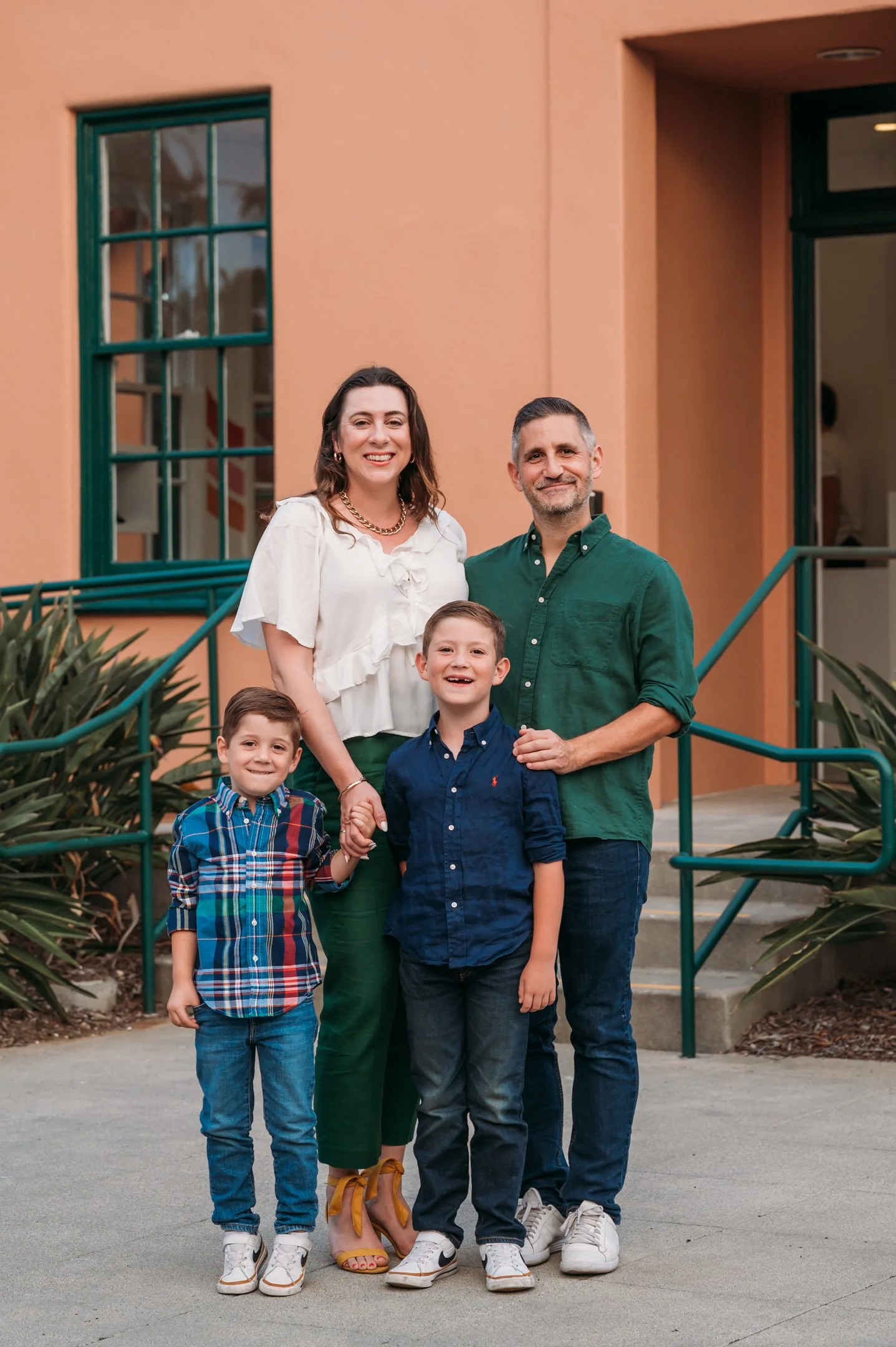 Family of four smiling outside a house with peach-colored walls and green windows, holding hands, with two children and two adults. Alisha Mowry Photography Military, Brand, and Portrait Photographer San Diego CA