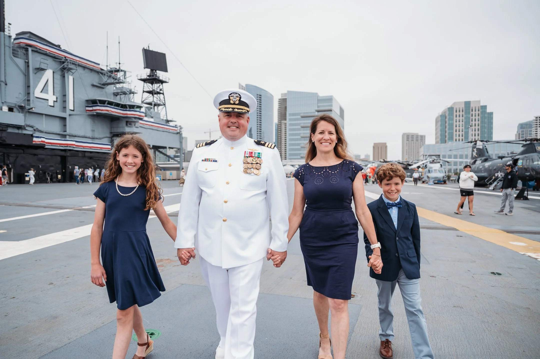 Family walking hand-in-hand on aircraft carrier deck, with a naval officer in uniform and children dressed formally, city skyline in background. Alisha Mowry Photography Military Photographer San Diego