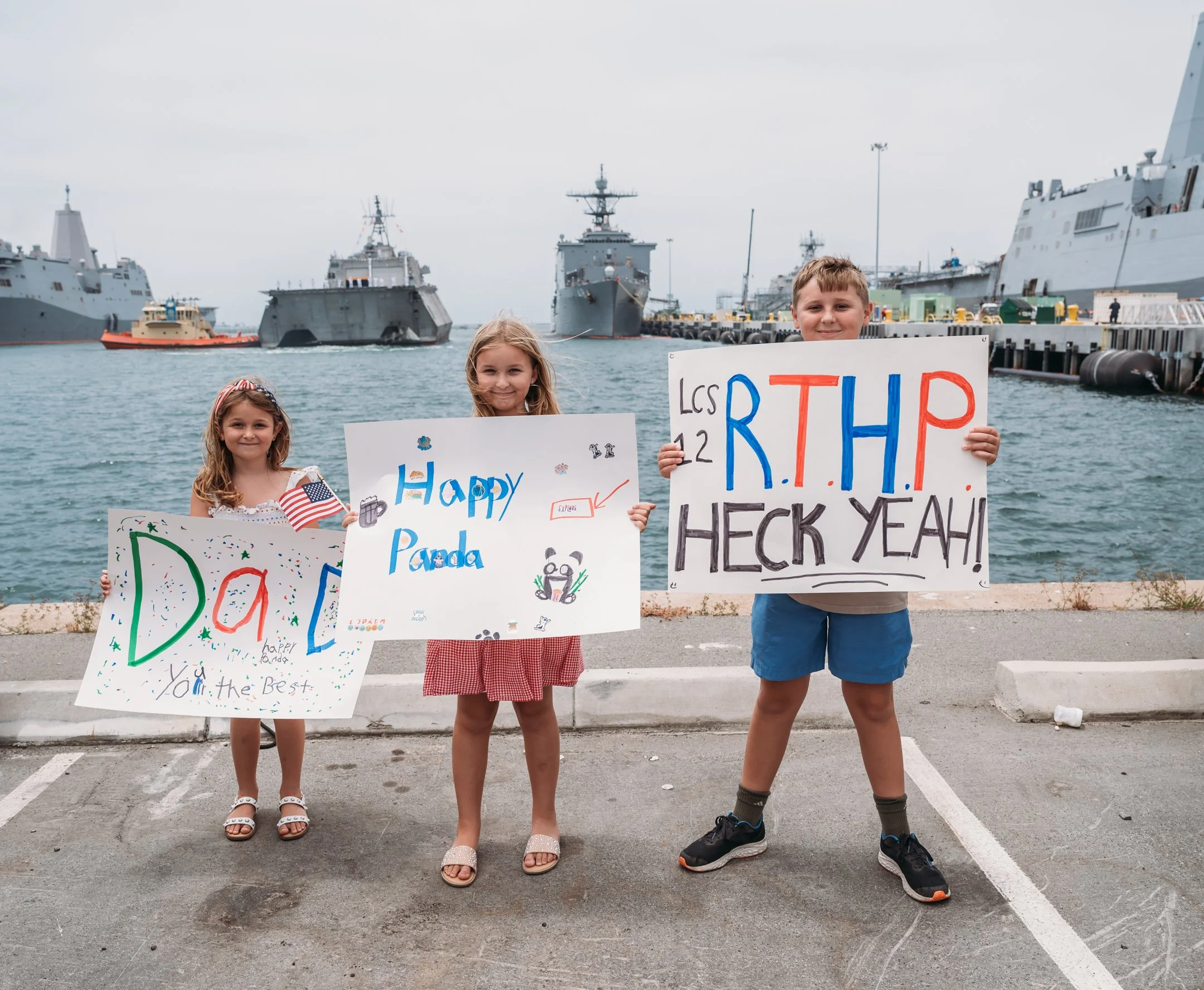Three children standing by a harbor holding colorful handmade signs celebrating a panda and the U.S. Navy, with ships in the background. Alisha Mowry Photography Military Photographer San Diego