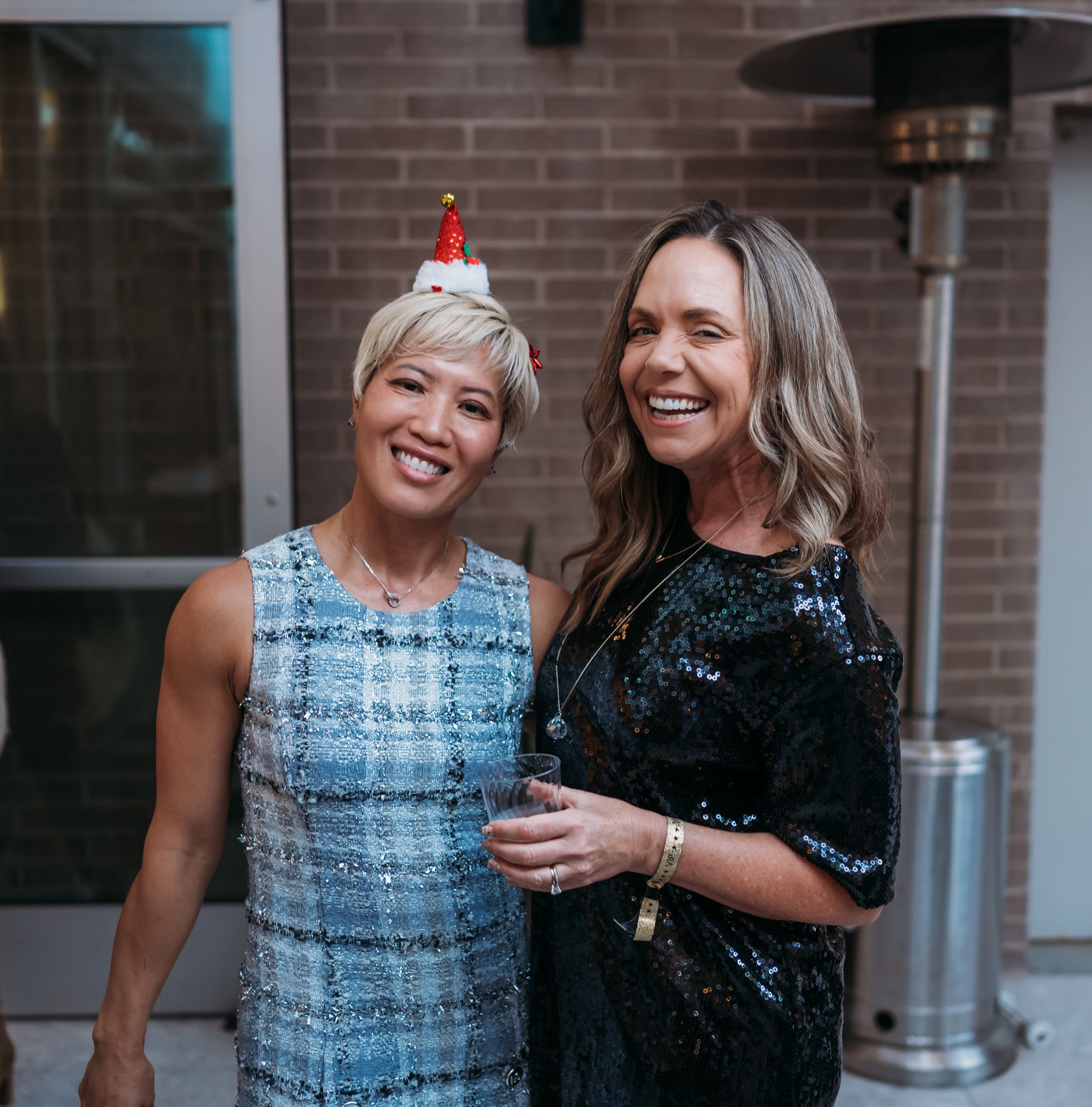 Two women at a party, smiling and dressed in shiny, festive outfits. One woman is wearing a sleeveless, blue patterned dress and a small red Christmas hat. The other woman is wearing a black sequin dress and holding a glass, with a big smile on her f