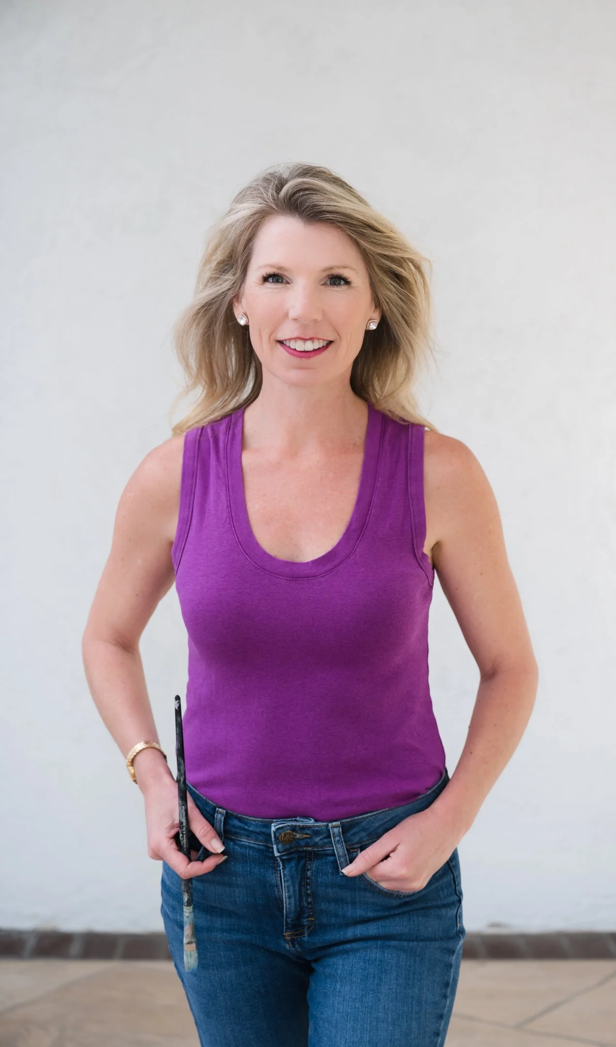 A woman with blonde, wavy hair, wearing a purple sleeveless top and blue jeans, standing against a neutral background, smiling and holding a paintbrush. Alisha Mowry Photography Military, Brand, and Portrait Photographer San Diego CA