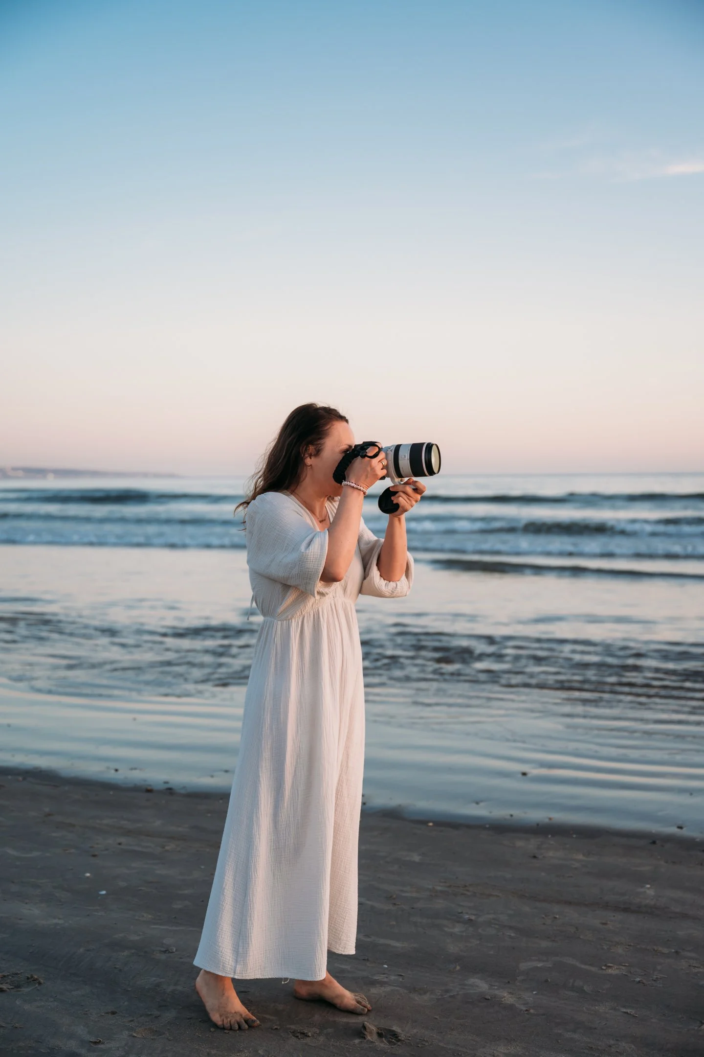 Woman in a white dress taking a photo with a camera on a beach at sunset. Alisha Mowry Photography Military, Brand, and Portrait Photographer San Diego CA