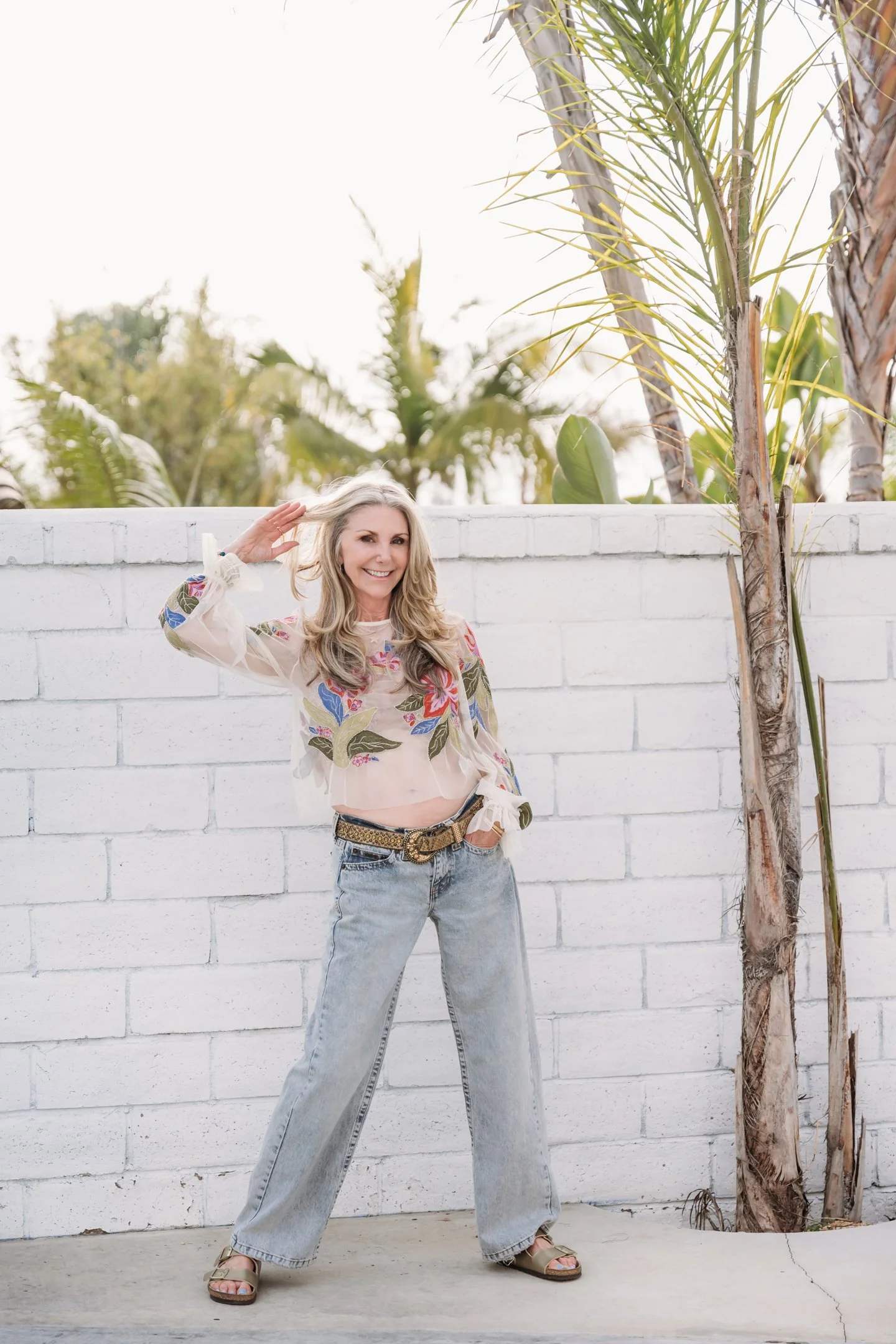 Woman with blonde hair wearing a floral embroidered long-sleeve shirt, blue jeans, and sandals standing against a white brick wall, smiling, with palm trees in the background. Alisha Mowry Photography Military, Brand, and Portrait Photographer San Di