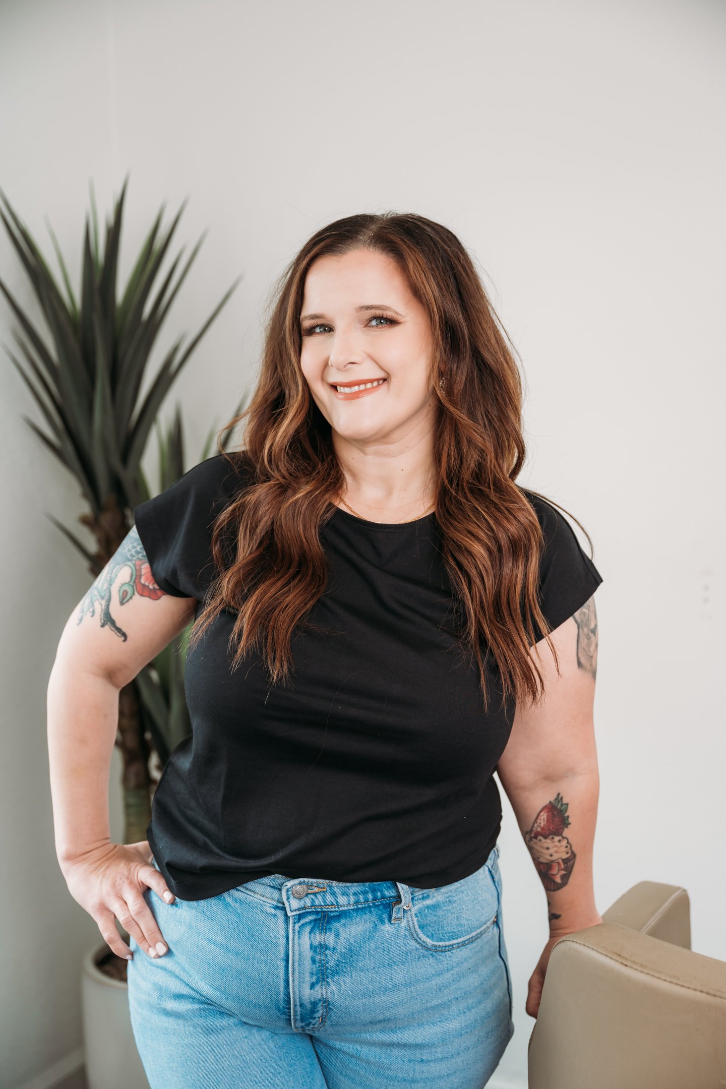 A woman with long wavy brown hair, wearing a black t-shirt and light blue jeans, smiling and standing indoors next to a beige chair and a tall green houseplant. Alisha Mowry Photography Military, Brand, and Portrait Photographer San Diego CA