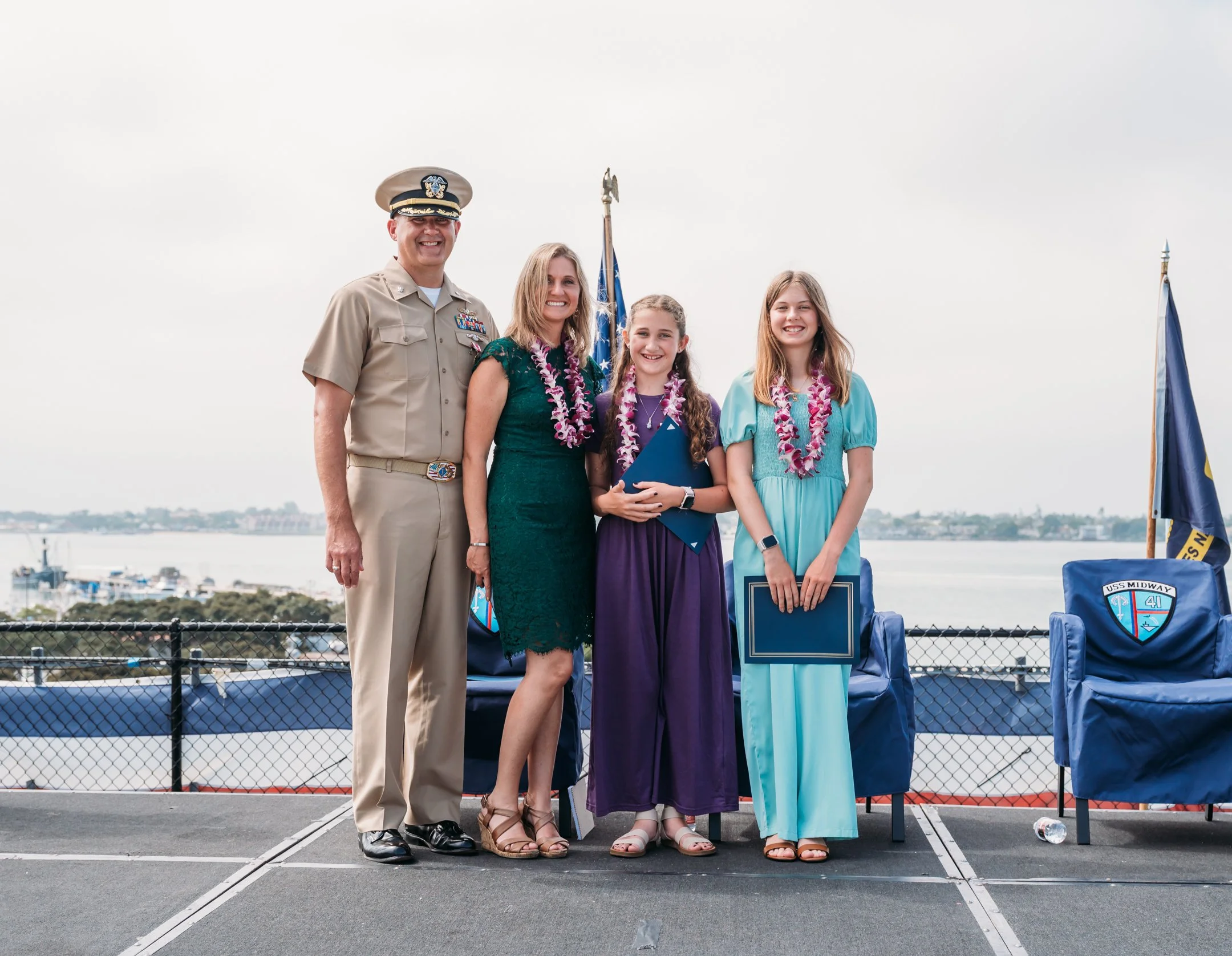 A military officer and two young girls standing outdoors at a celebration, wearing leis, holding certificates and folders, with a body of water and boats in the background. uss midway military retirement ceremony 