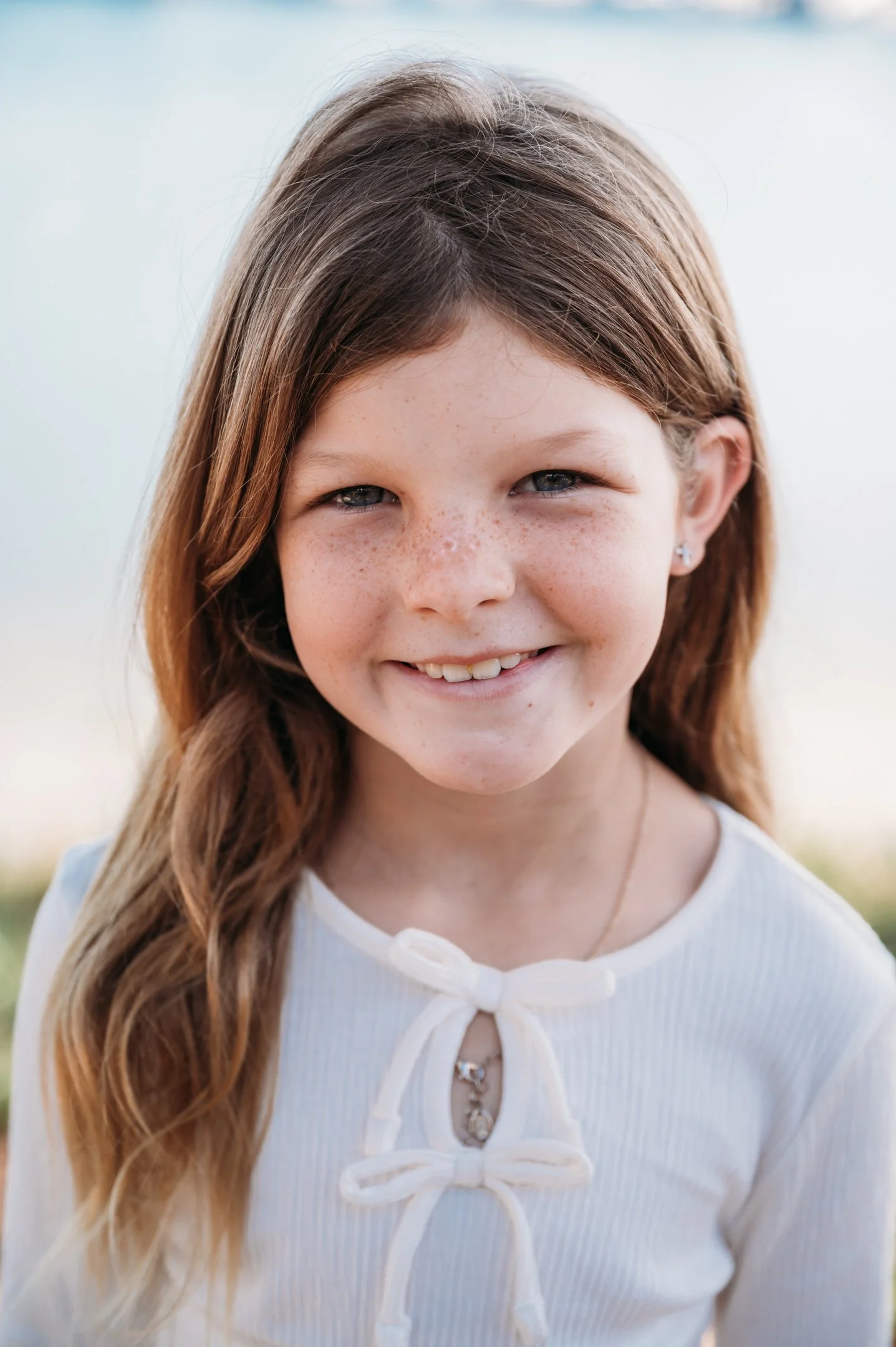 Close-up of a smiling young girl with brown hair and freckles, wearing a white top with bows and a necklace, outdoors with a blurred background. Alisha Mowry Photography Military, Brand, and Portrait Photographer San Diego CA