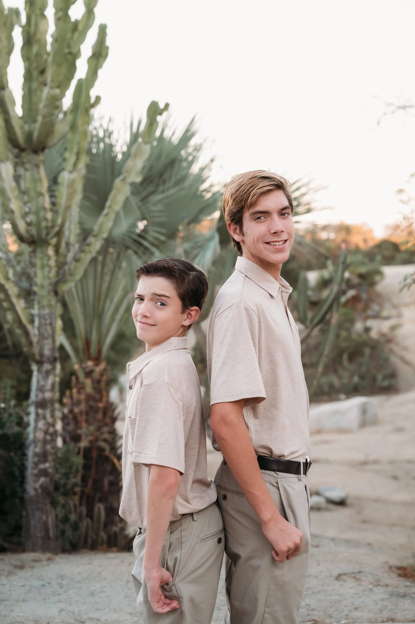 Two boys standing back-to-back outdoors in a desert landscape with large cacti in the background, both wearing beige shirts and khaki pants, smiling with relaxed expressions. Alisha Mowry Photography Military, Brand, and Portrait Photographer San Die