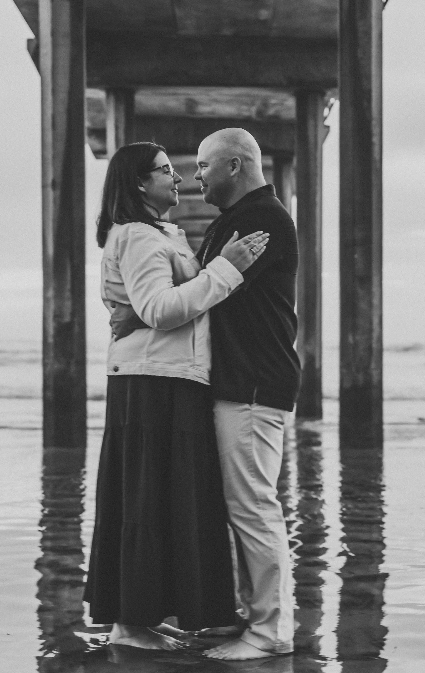 A couple embracing on a beach under a pier, facing each other and smiling, with the water and sky in the background.