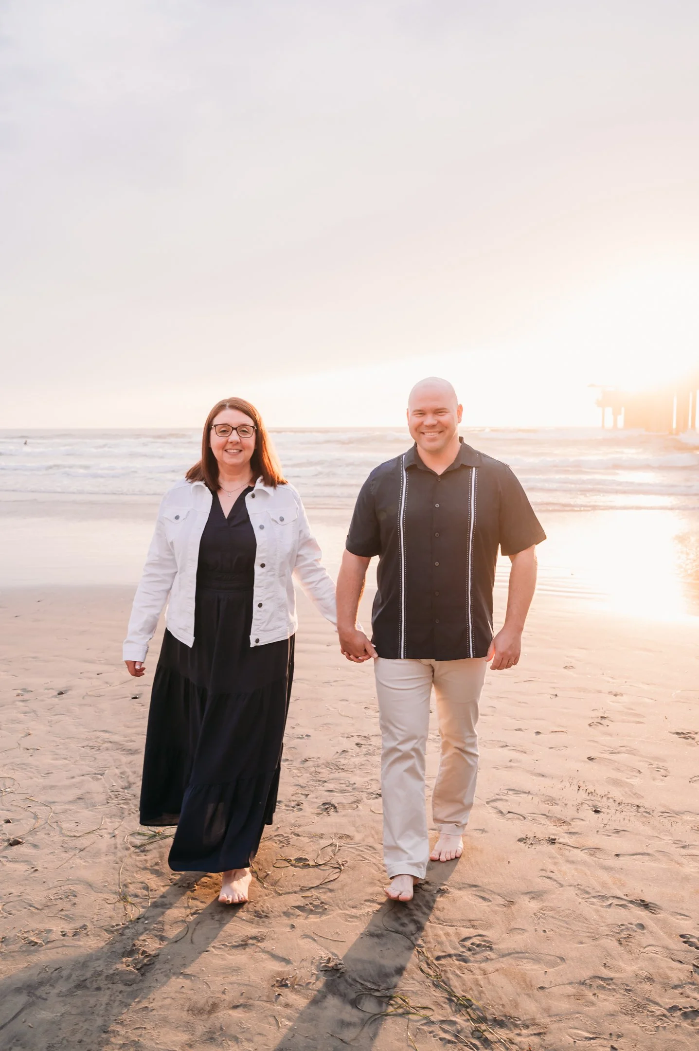 A smiling couple walking hand in hand on the beach during sunset, with the ocean in the background.