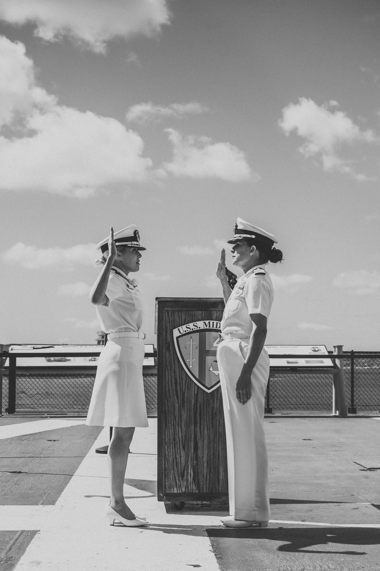 Two women in naval uniforms exchanging hand salutes in front of a wood podium with a shield emblem reading 'U.S.S. MIDWAY' on a naval ship deck.  Alisha Mowry Photography Military Photographer San Diego