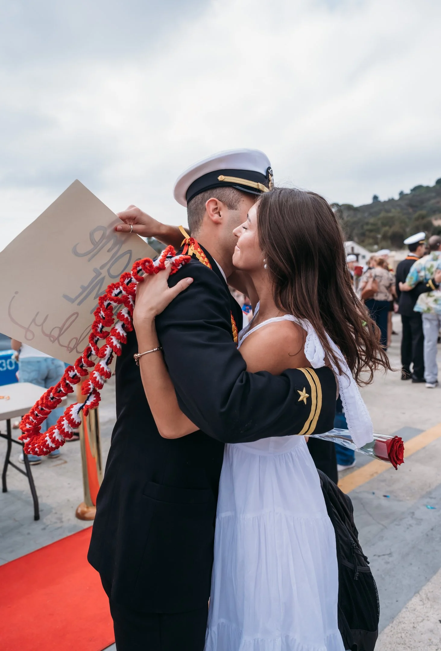 A woman and a man in military uniform embrace each other during a graduation ceremony, with the woman holding a single rose and the man wearing a garland and holding a sign. Alisha Mowry Photography Military Photographer San Diego