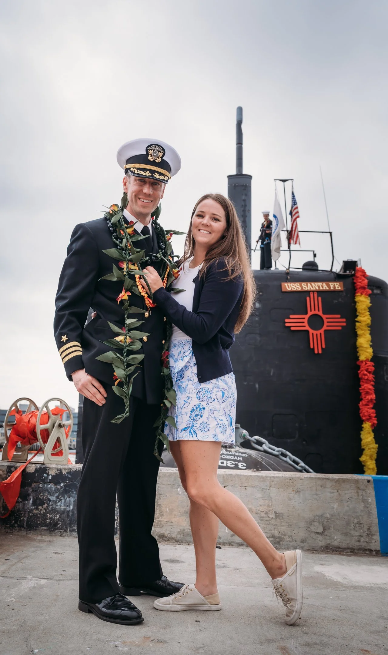 A man in a navy military uniform and a woman in casual clothes stand close together, smiling, in front of a navy submarine decorated with flowers, flags, and a sign reading 'USS Santa Fe'. Alisha Mowry Photography Military Photographer San Diego